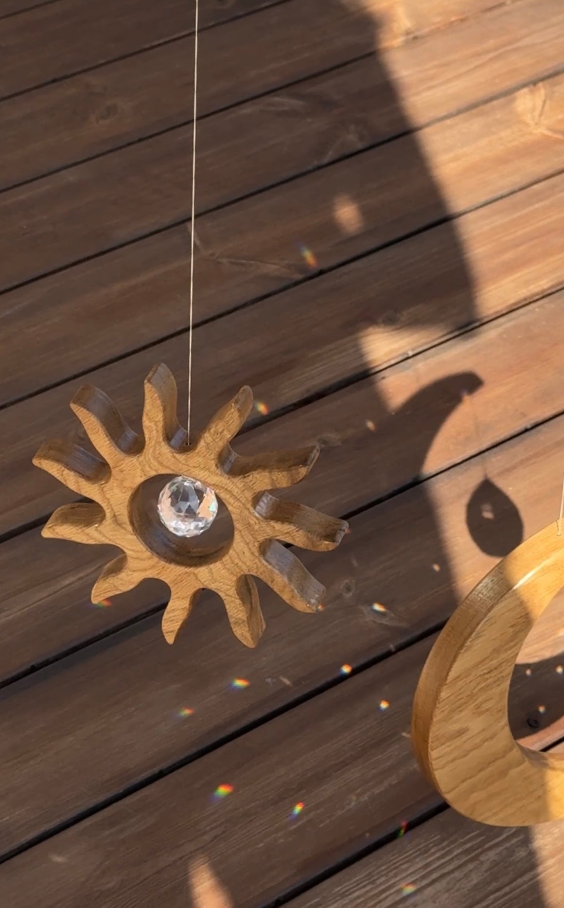 Handgefertigter Sonnenfänger aus Eichenholz (Oak) in Sonnenform with a central Asfour crystal prism. The Solstice wooden suncatcher casting rainbows on a white wall. Hochwertige Wohndekoration und Regenbogenmacher für Fenster oder Balkon.