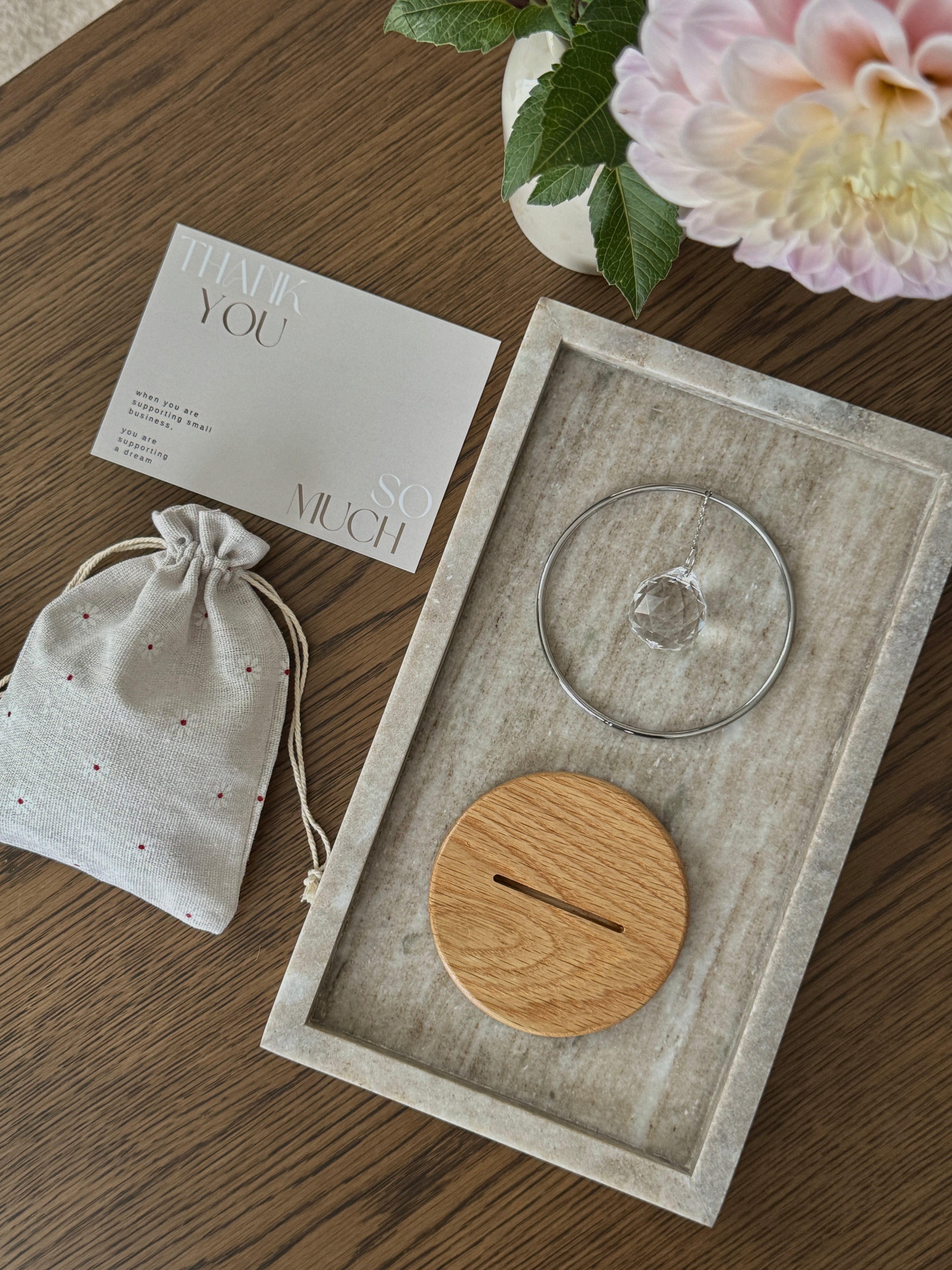 Decorative items including a wooden coaster, glass lid, and gift bag on a wooden surface with flowers in the background.