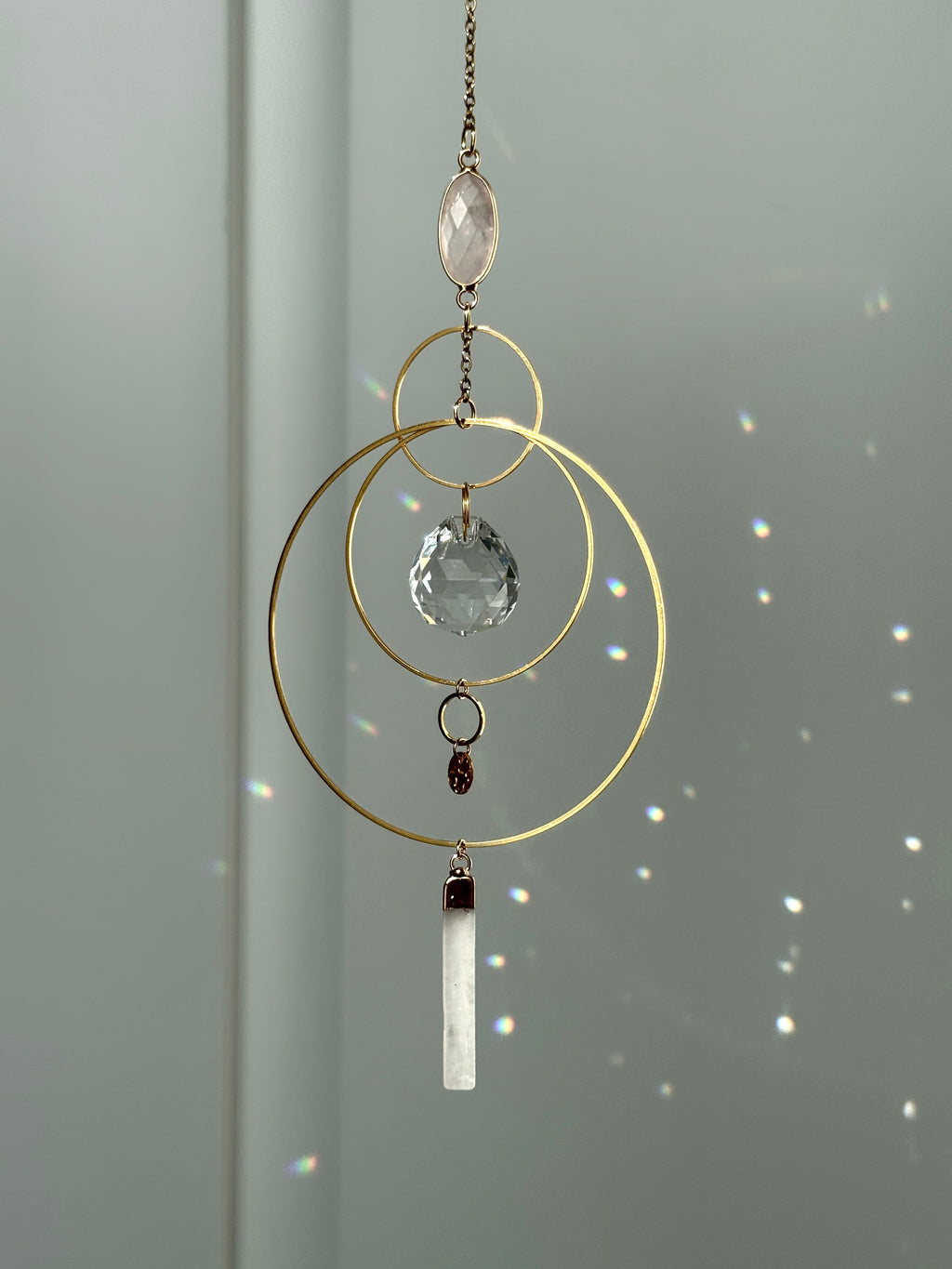 A gold geometric suncatcher featuring two pink Rose Quartz gemstones and a faceted crystal sphere. The suncatcher consists of two interlocking gold hoops, a textured coin charm, and is hanging against a soft-focus background, reflecting bright light.