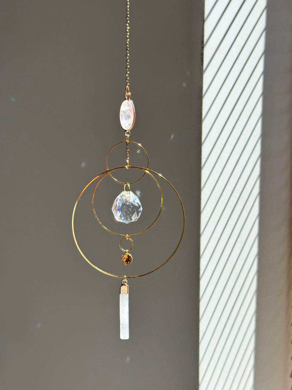 A gold geometric suncatcher featuring two pink Rose Quartz gemstones and a faceted crystal sphere. The suncatcher consists of two interlocking gold hoops, a textured coin charm, and is hanging against a soft-focus background, reflecting bright light.