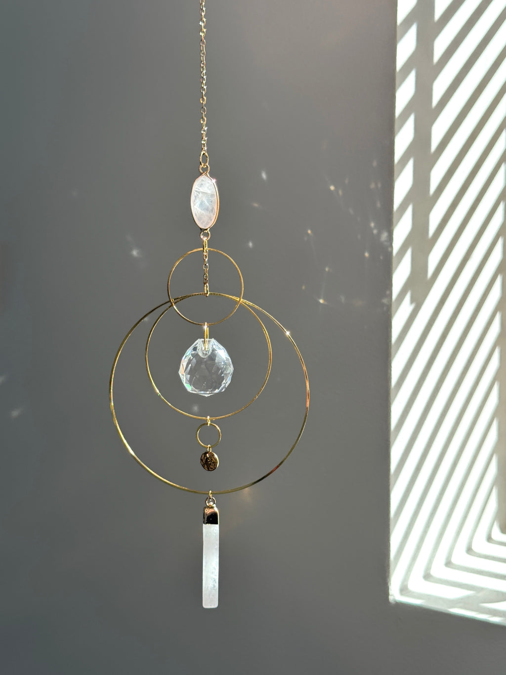 A gold geometric suncatcher featuring two pink Rose Quartz gemstones and a faceted crystal sphere. The suncatcher consists of two interlocking gold hoops, a textured coin charm, and is hanging against a soft-focus background, reflecting bright light.