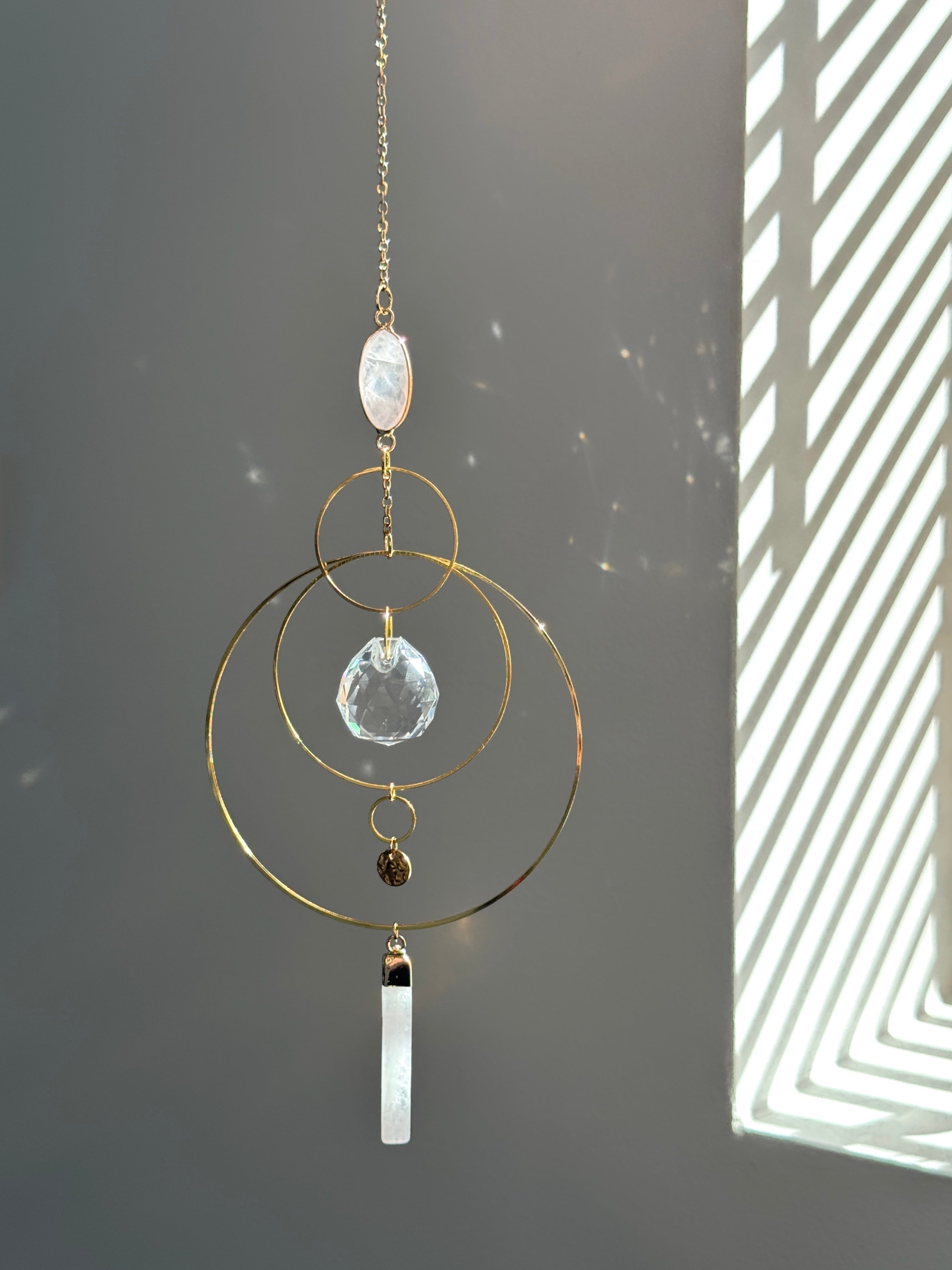 A gold geometric suncatcher featuring two pink Rose Quartz gemstones and a faceted crystal sphere. The suncatcher consists of two interlocking gold hoops, a textured coin charm, and is hanging against a soft-focus background, reflecting bright light.