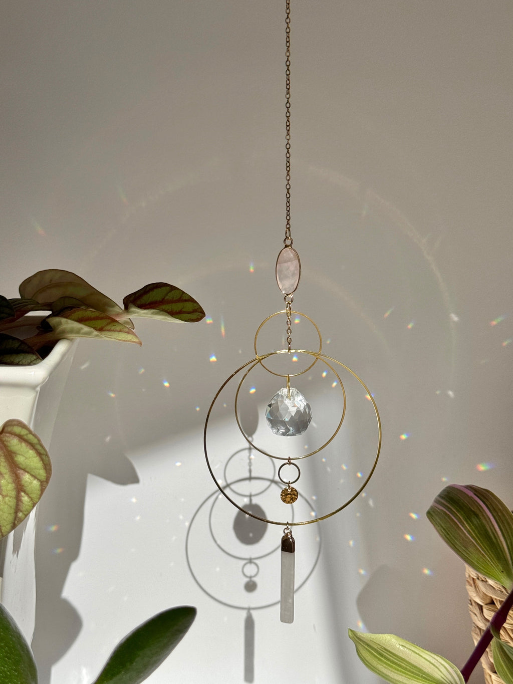 A gold geometric suncatcher featuring two pink Rose Quartz gemstones and a faceted crystal sphere. The suncatcher consists of two interlocking gold hoops, a textured coin charm, and is hanging against a soft-focus background, reflecting bright light.
