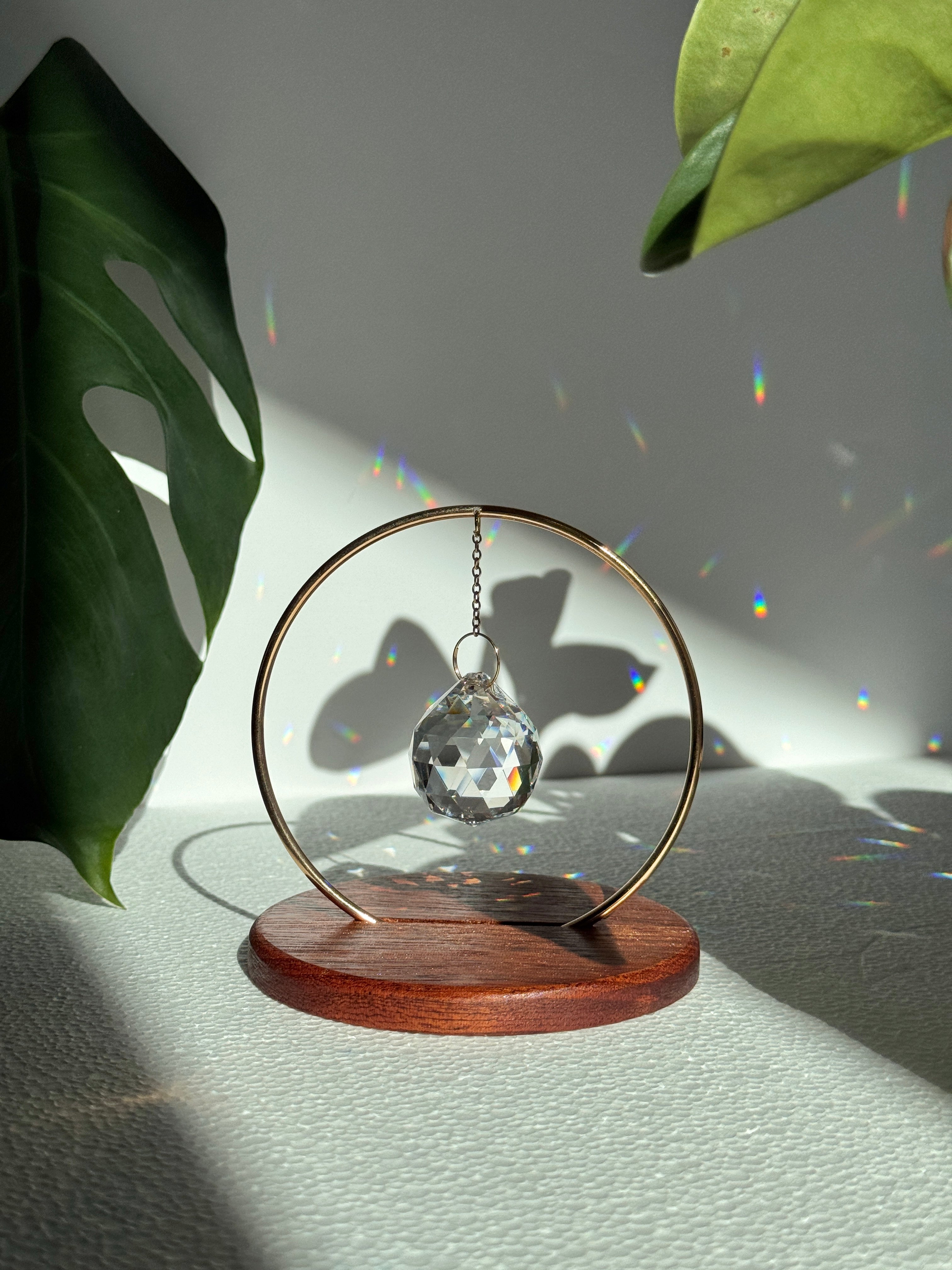 The Circle - A minimalist Sonnenfänger (suncatcher) featuring a high-quality faceted Kristall-Prisma suspended by a delicate gold stainless steel chain within a matching gold ring. The modern tabletop design rests on a solid, dark redwood base (Echtholz-Sockel) and is shown creating vibrant rainbow light reflections (Regenbogen-Effekt) across the background. This elegant Fensterdeko is perfect as a mindful Geschenkidee for home or office.