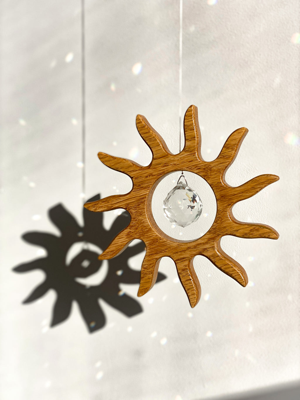 Handgefertigter Sonnenfänger aus Eichenholz (Oak) in Sonnenform with a central Asfour crystal prism. The Solstice wooden suncatcher casting rainbows on a white wall. Hochwertige Wohndekoration und Regenbogenmacher für Fenster oder Balkon.