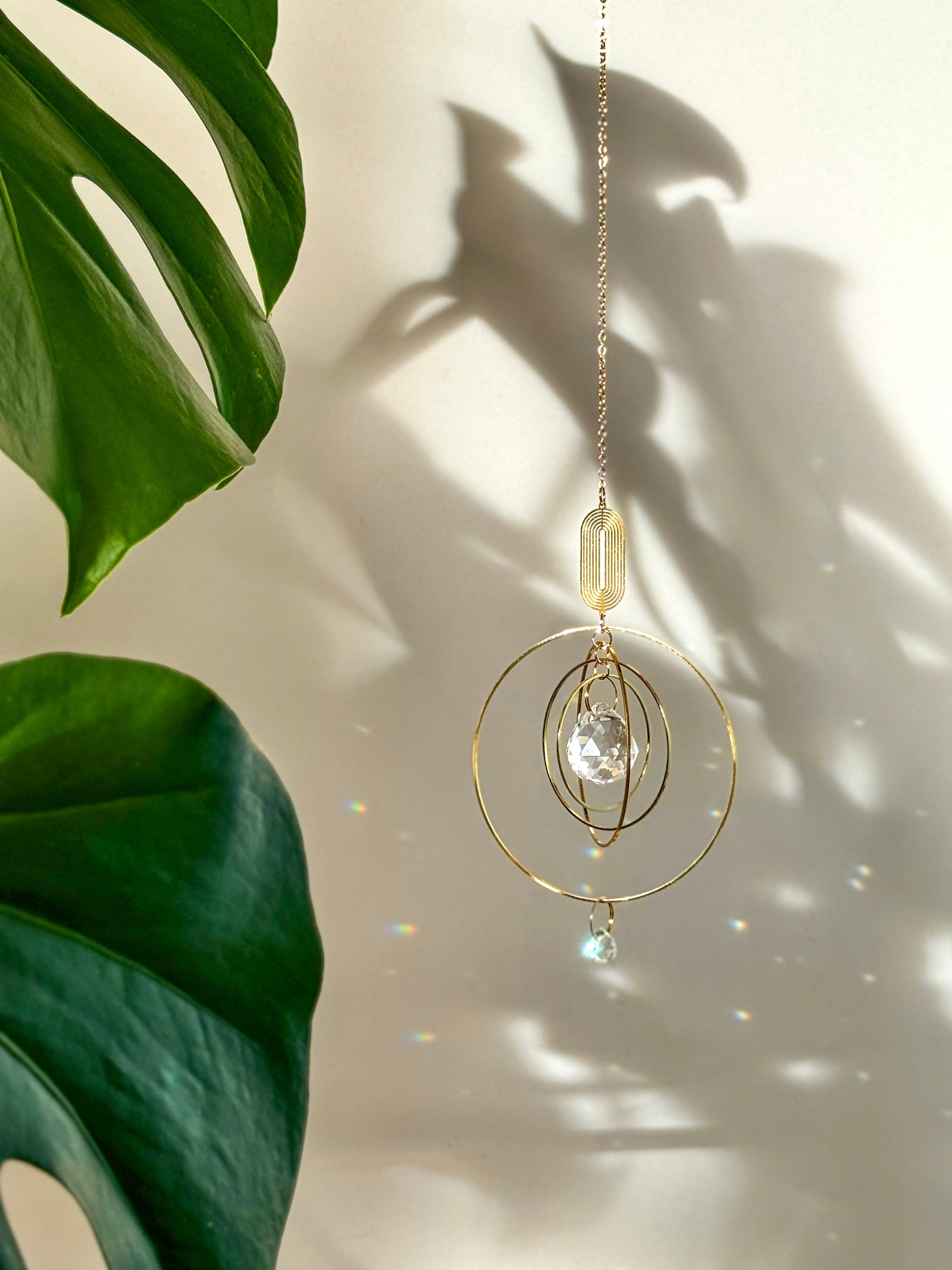 Celestial boho window suncatcher (Sonnenfänger Fenster) with gold concentric rings and a crystal prism, scattering magical rainbows (Regenbogen) across a sunlit wall next to a Monstera leaf.