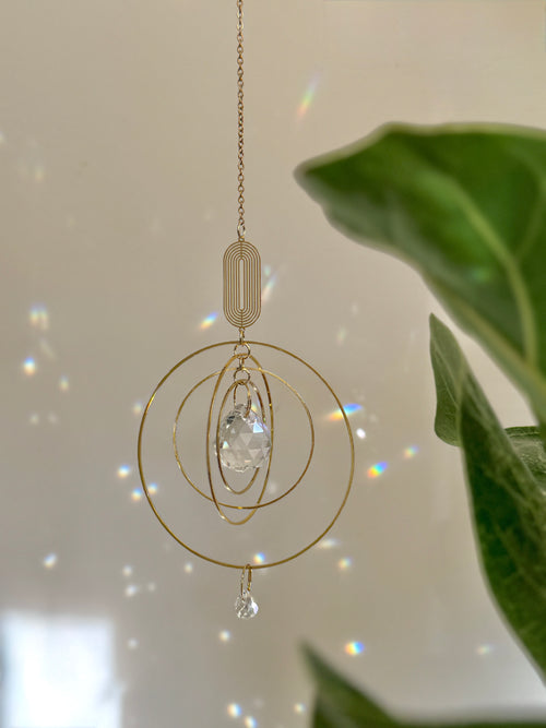 Celestial boho window suncatcher (Sonnenfänger Fenster) with gold concentric rings and a crystal prism, scattering magical rainbows (Regenbogen) across a sunlit wall next to a Monstera leaf.