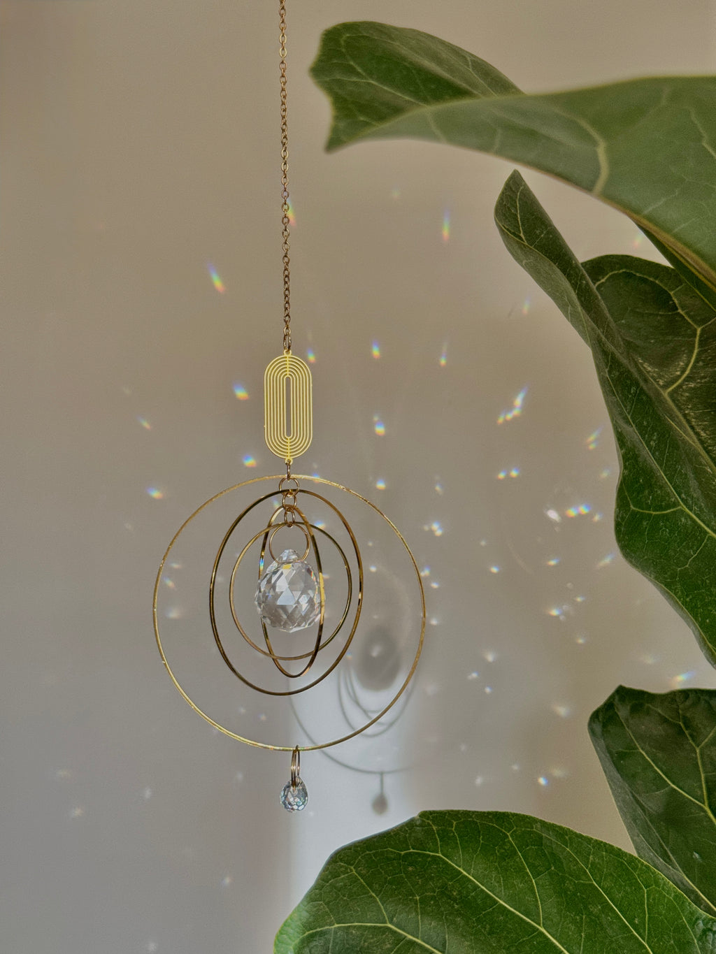 Celestial boho window suncatcher (Sonnenfänger Fenster) with gold concentric rings and a crystal prism, scattering magical rainbows (Regenbogen) across a sunlit wall next to a Monstera leaf.