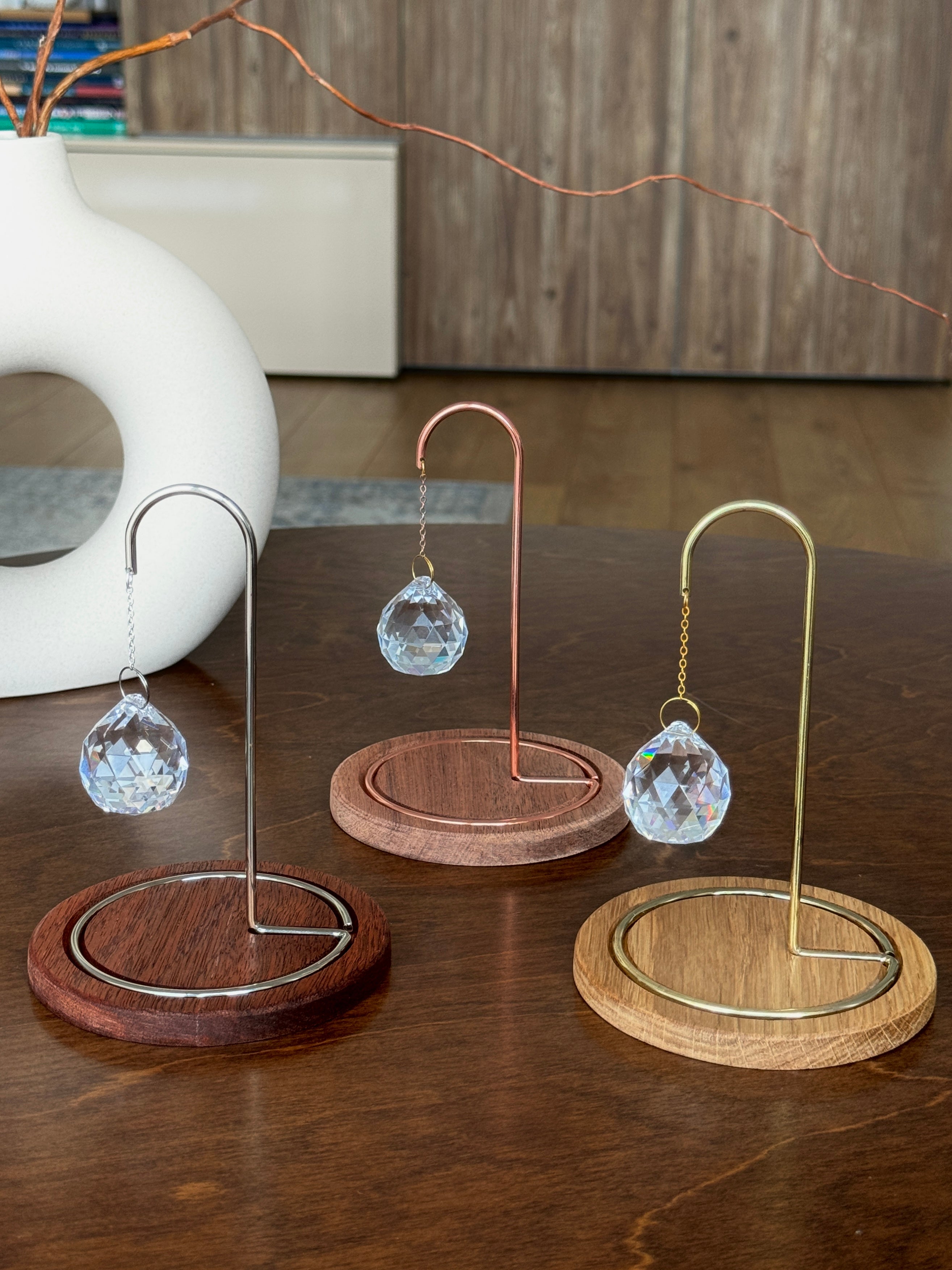 A faceted crystal suncatcher hangs from a rose gold metal hook on a circular wooden base. Sunlight streams through the crystal, scattering vibrant rainbow-colored light reflections across the base and the surrounding white surface. The wooden base is engraved with the message: "You are your home. Take care of yourself." followed by a small hand-drawn heart.