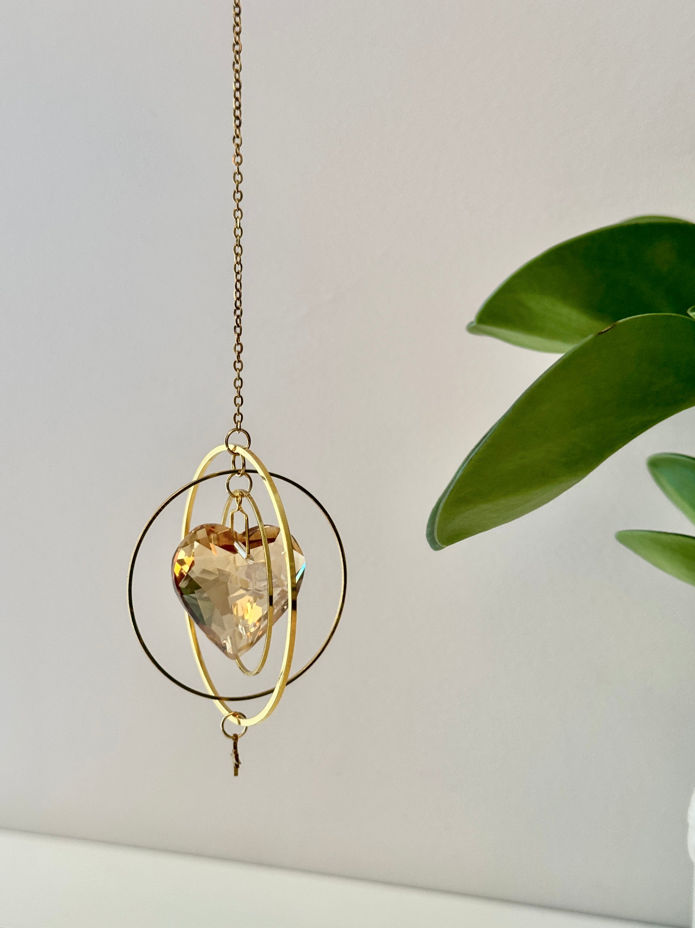 Sunkissed Heartbeat crystal suncatcher featuring a warm amber-gold faceted heart prism. Handmade celestial window decor with gold rings casting rainbow light flares (Sonnenfänger).