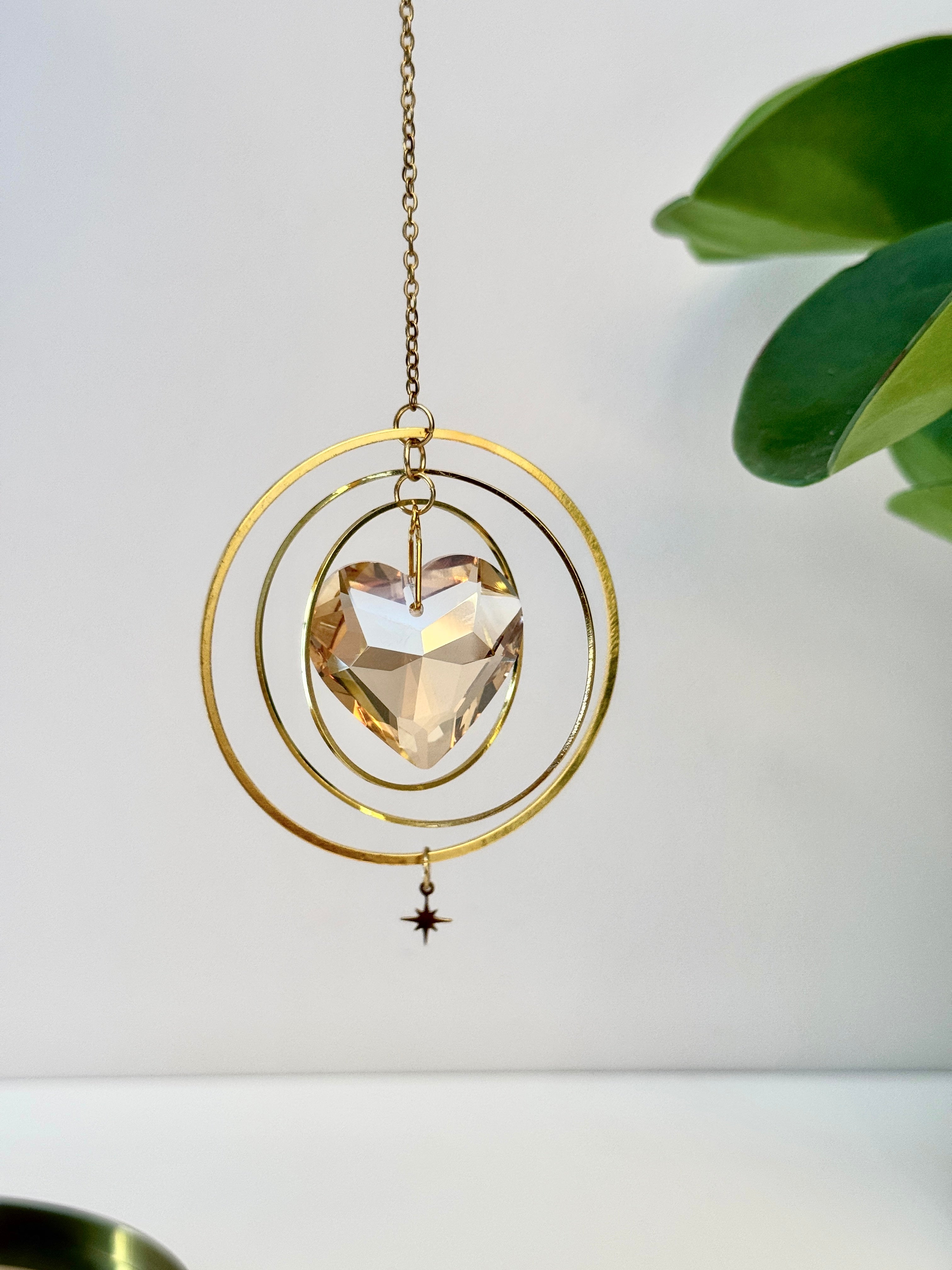 Sunkissed Heartbeat crystal suncatcher featuring a warm amber-gold faceted heart prism. Handmade celestial window decor with gold rings casting rainbow light flares (Sonnenfänger).