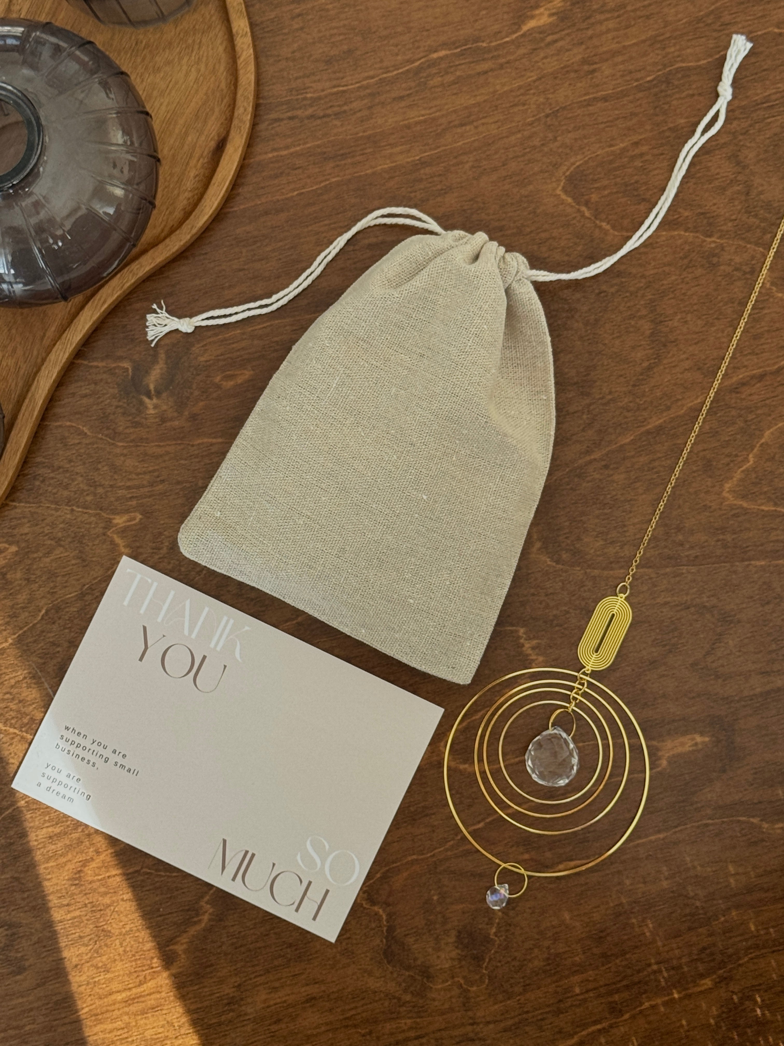Gift-ready suncatcher (Sonnenfänger Geschenk) featuring a golden celestial prism alongside a linen drawstring bag and a "Thank You" card on a wooden surface.