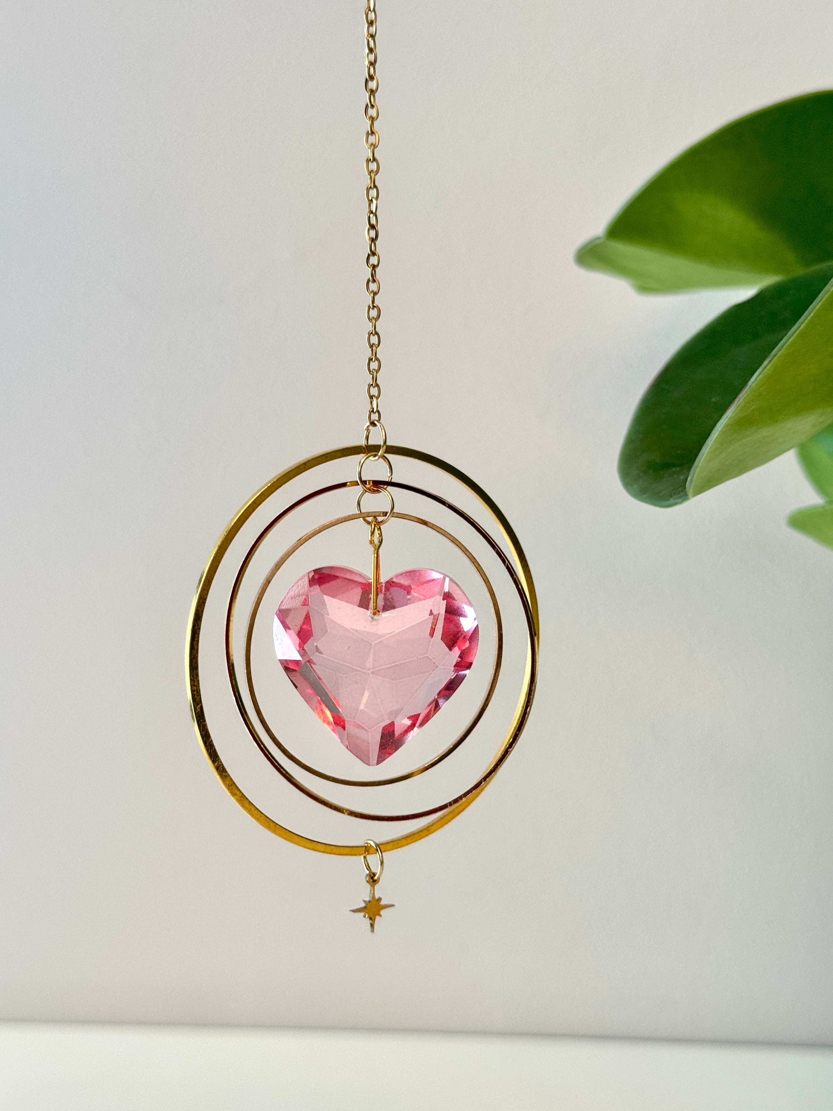 Sunkissed Heartbeat crystal suncatcher featuring a pink-gold faceted heart prism. Handmade celestial window decor with gold rings casting rainbow light flares (Sonnenfänger).