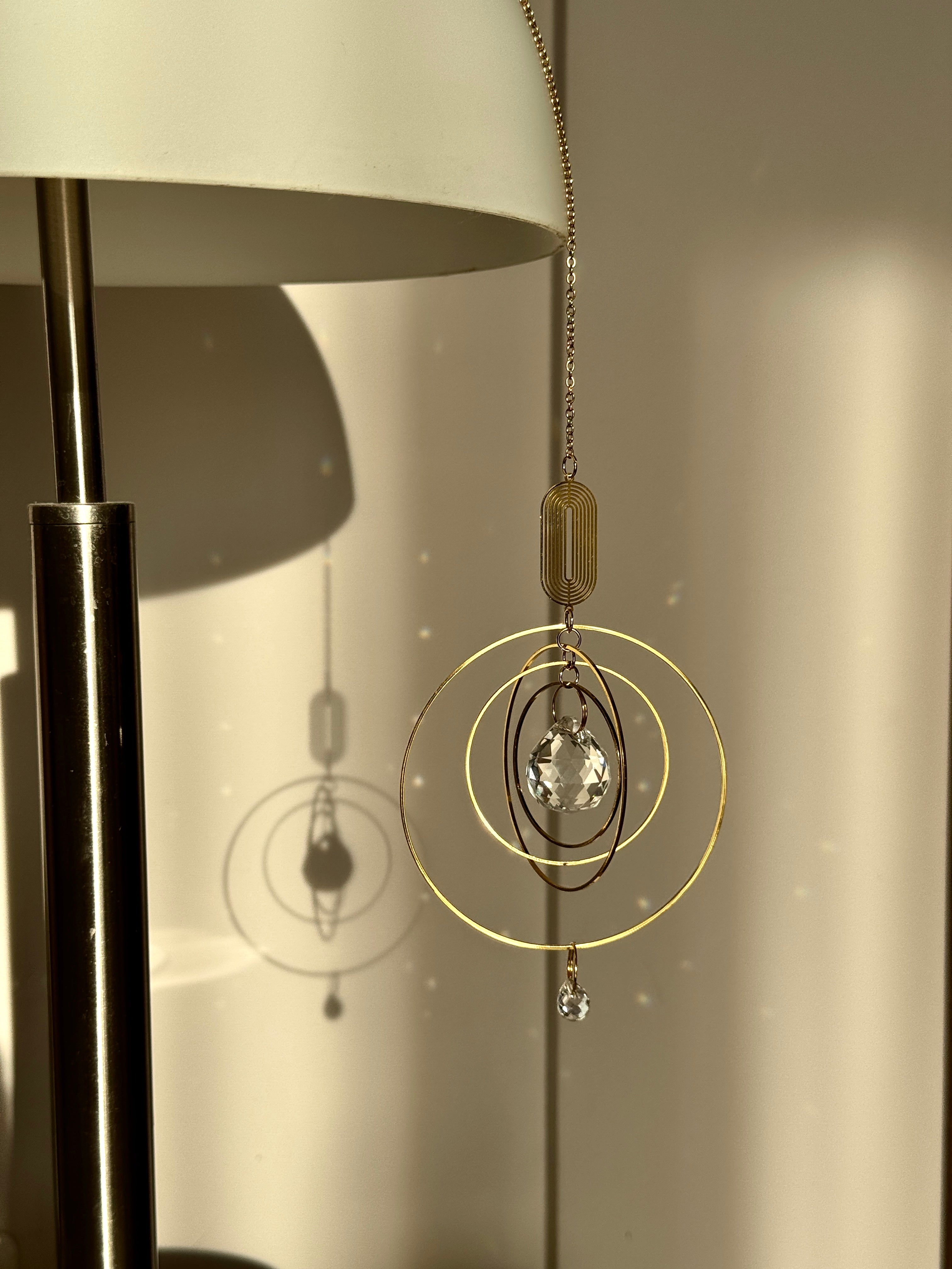 Golden boho suncatcher (Fensterdeko) hanging near an indoor lamp, showing how the crystal prism (Kristall) creates soft light reflections even in low evening light.