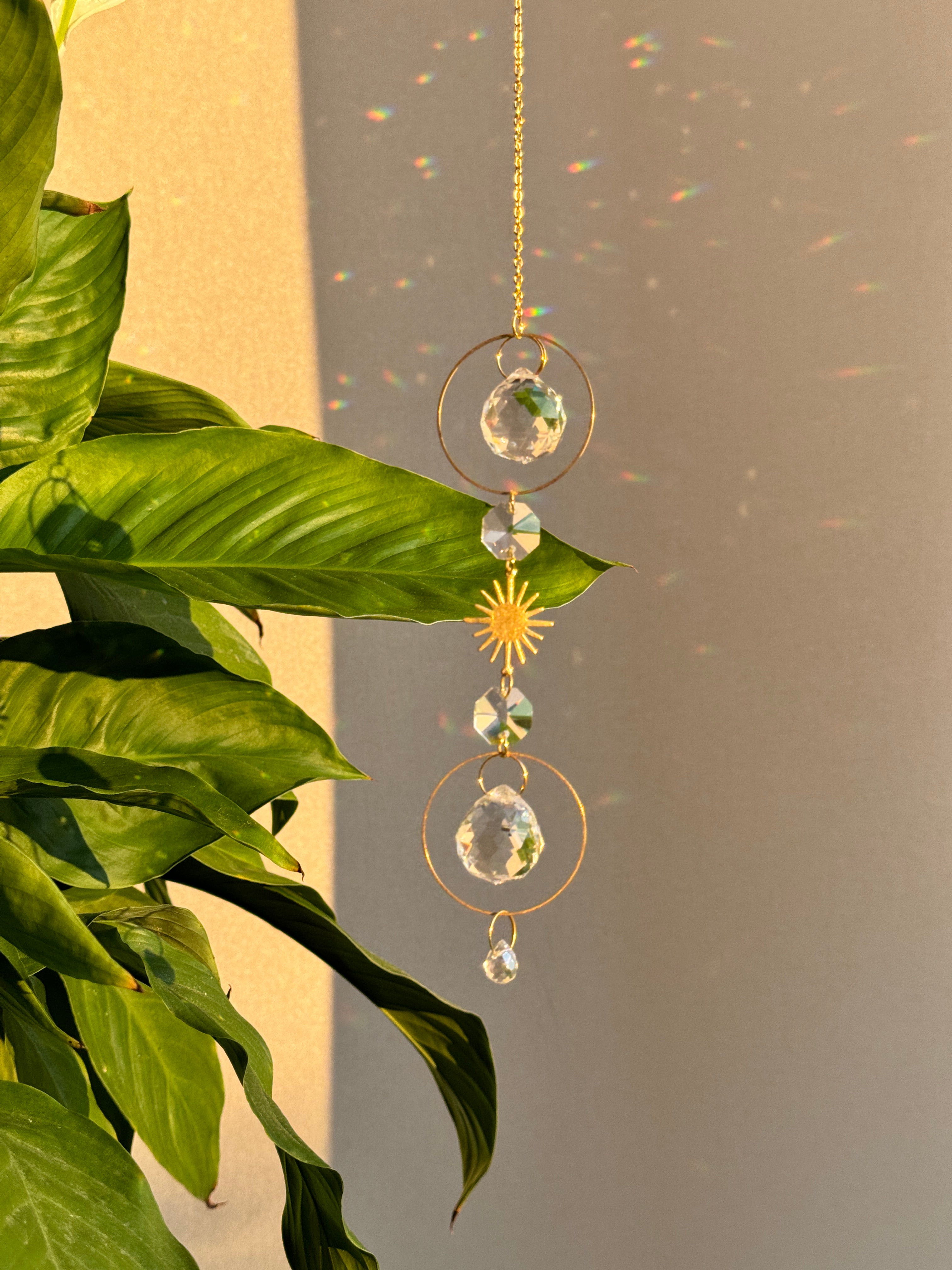 Bestselling Golden Hour suncatcher hanging indoors, featuring gold geometric rings, faceted crystal spheres, and a starburst charm. Dieser handgefertigte Sonnenfänger casts brilliant rainbow patterns and Lichtreflexe against a warm wall. Perfekte Boho-Stil Fensterdeko.