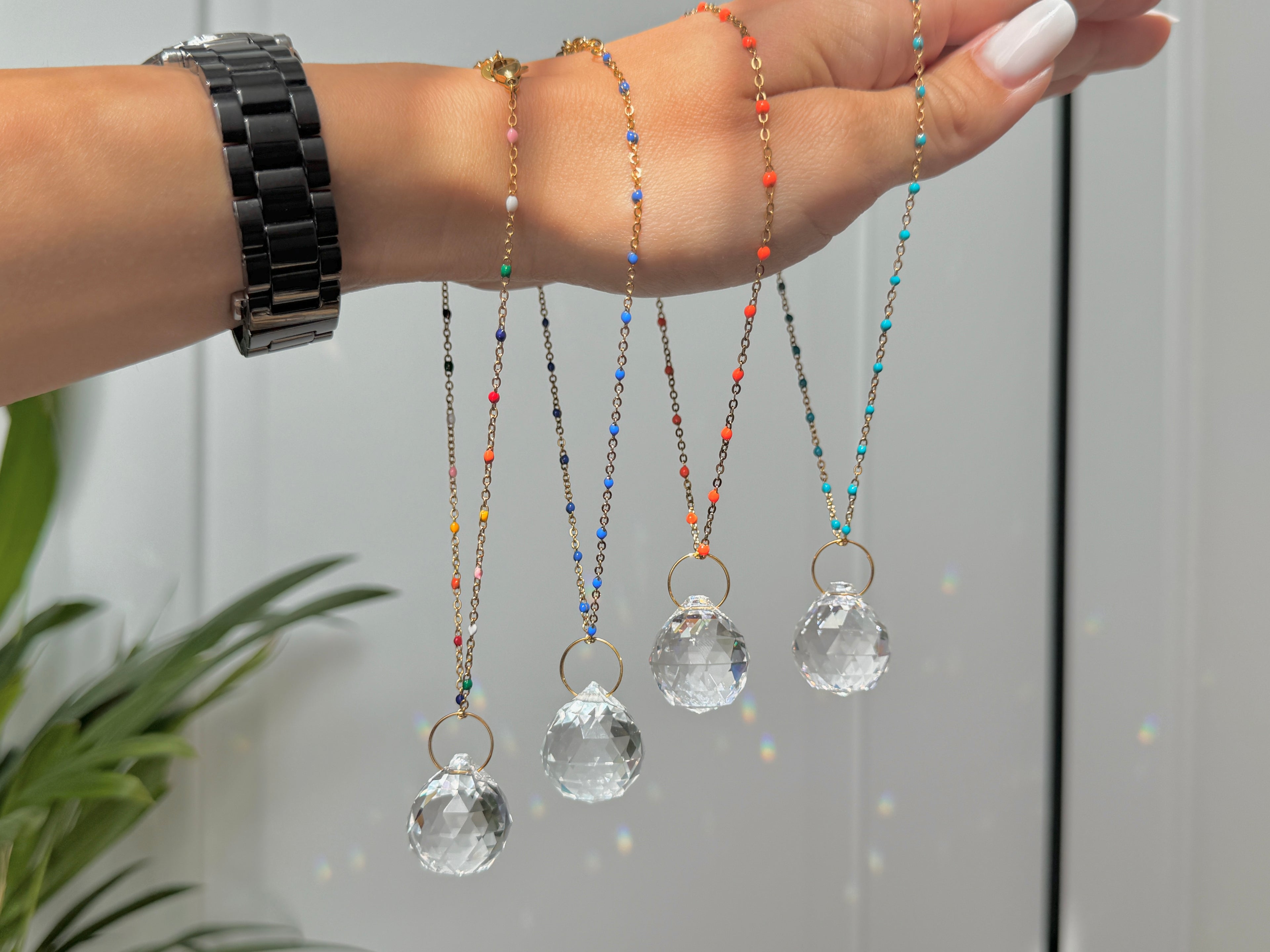 Fourcrystal prism car suncatchers in different sizes hanging from a hand, casting rainbow light reflections on a white wall next to a green plant.