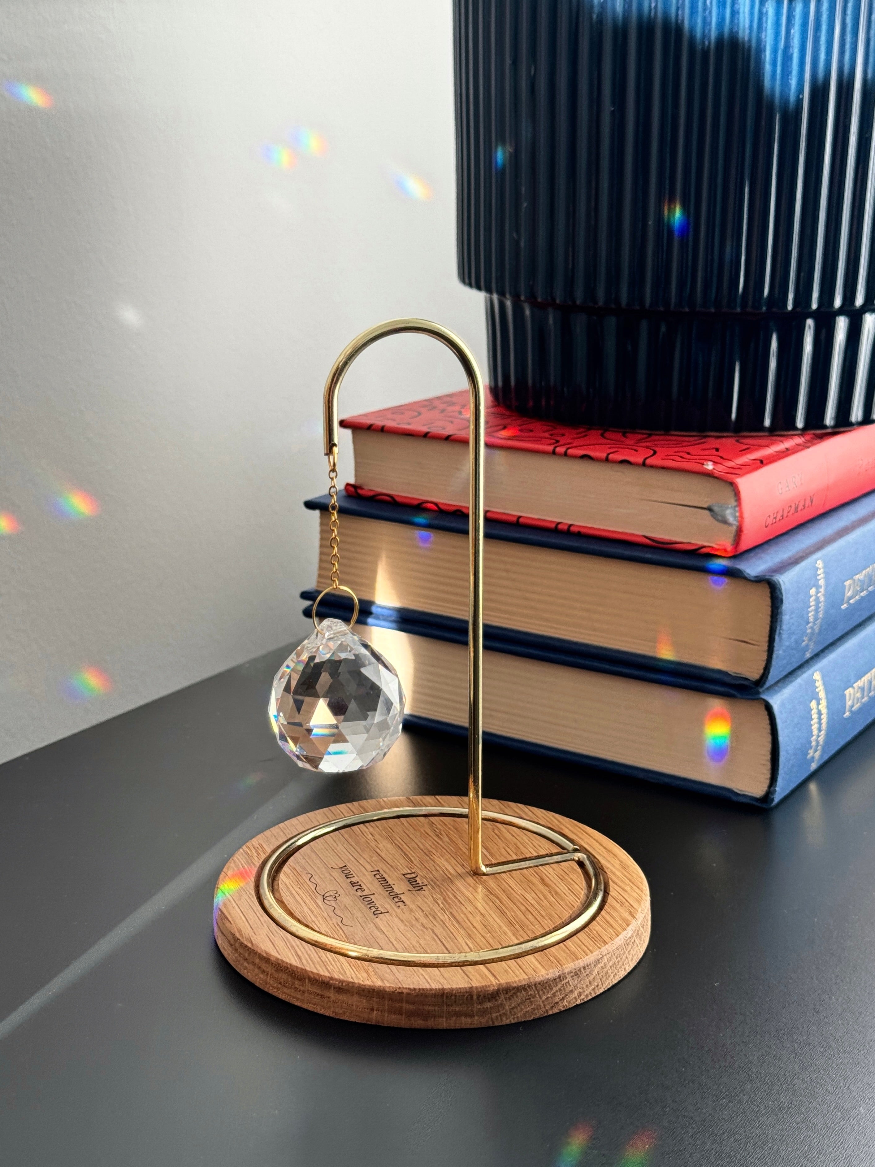 A faceted glass crystal sphere hangs from a gold wire stand on a circular wooden base, which is engraved with the words "Daily reminder: you are loved." The suncatcher is on a white surface next to a wooden window blind. Sunlight streams through the crystal, creating a radiant glow and casting intricate patterns of light and rainbow-colored reflections on the base and surrounding surfaces.