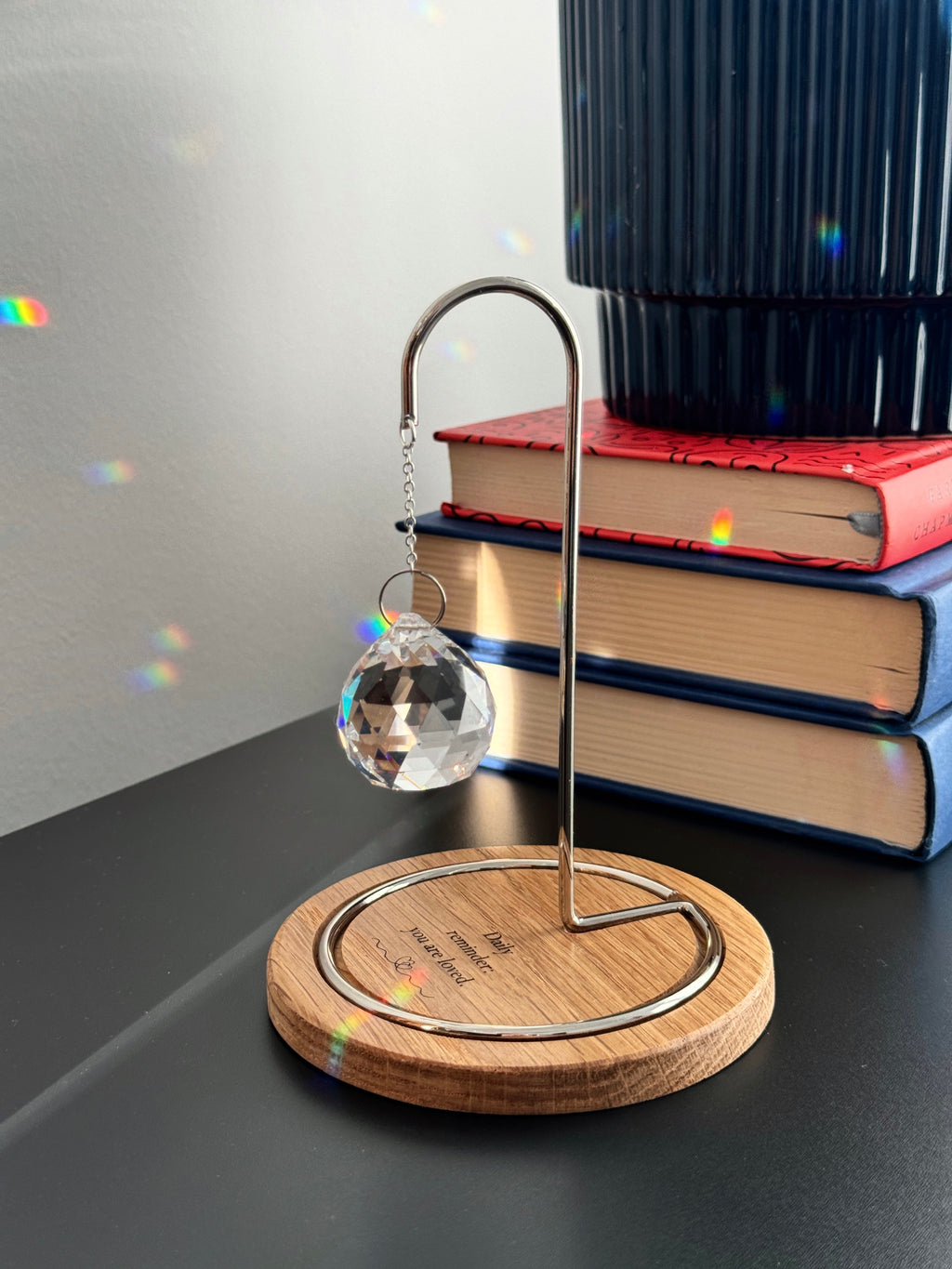 A faceted glass crystal sphere hangs from a gold wire stand on a circular wooden base, which is engraved with the words "Daily reminder: you are loved." The suncatcher is on a white surface next to a wooden window blind. Sunlight streams through the crystal, creating a radiant glow and casting intricate patterns of light and rainbow-colored reflections on the base and surrounding surfaces.