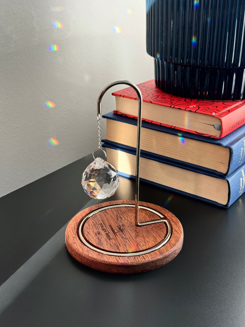 A faceted glass crystal sphere hangs from a gold wire stand on a circular wooden base, which is engraved with the words "Daily reminder: you are loved." The suncatcher is on a white surface next to a wooden window blind. Sunlight streams through the crystal, creating a radiant glow and casting intricate patterns of light and rainbow-colored reflections on the base and surrounding surfaces.