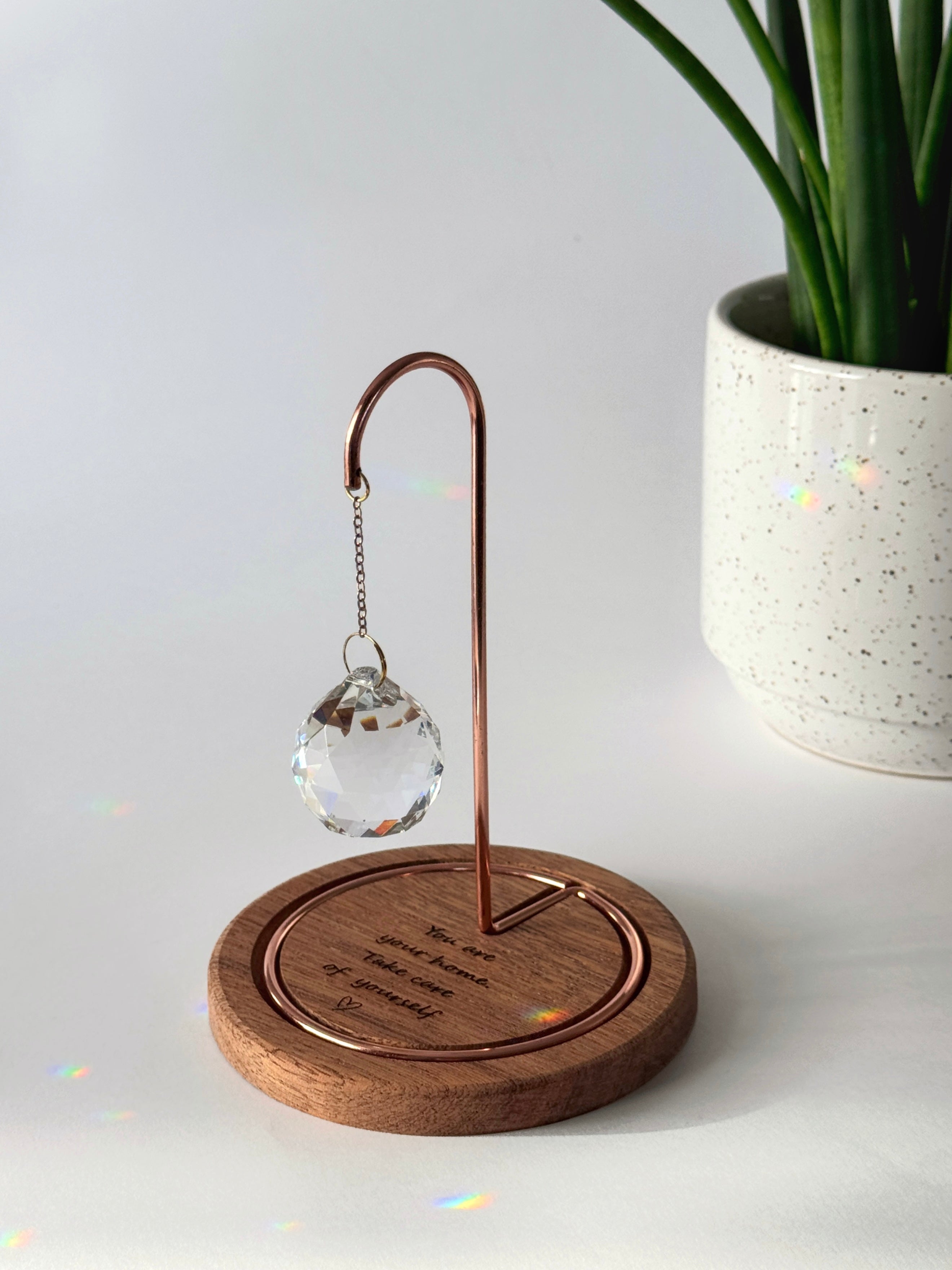 A faceted crystal suncatcher hangs from a rose gold metal hook on a circular wooden base. Sunlight streams through the crystal, scattering vibrant rainbow-colored light reflections across the base and the surrounding white surface. The wooden base is engraved with the message: "You are your home. Take care of yourself." followed by a small hand-drawn heart.