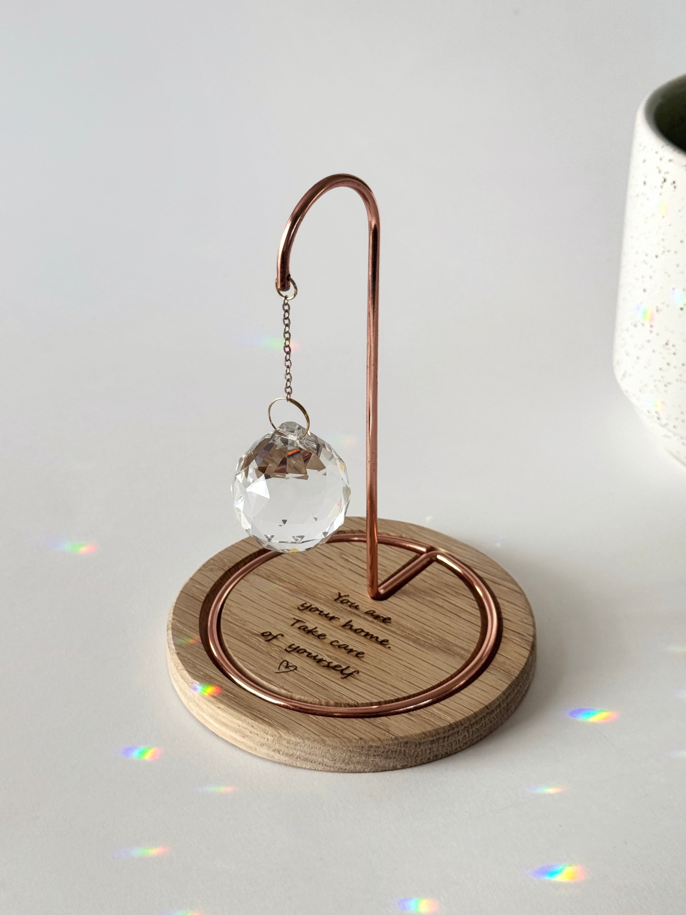 A faceted crystal suncatcher hangs from a rose gold metal hook on a circular wooden base. Sunlight streams through the crystal, scattering vibrant rainbow-colored light reflections across the base and the surrounding white surface. The wooden base is engraved with the message: "You are your home. Take care of yourself." followed by a small hand-drawn heart.