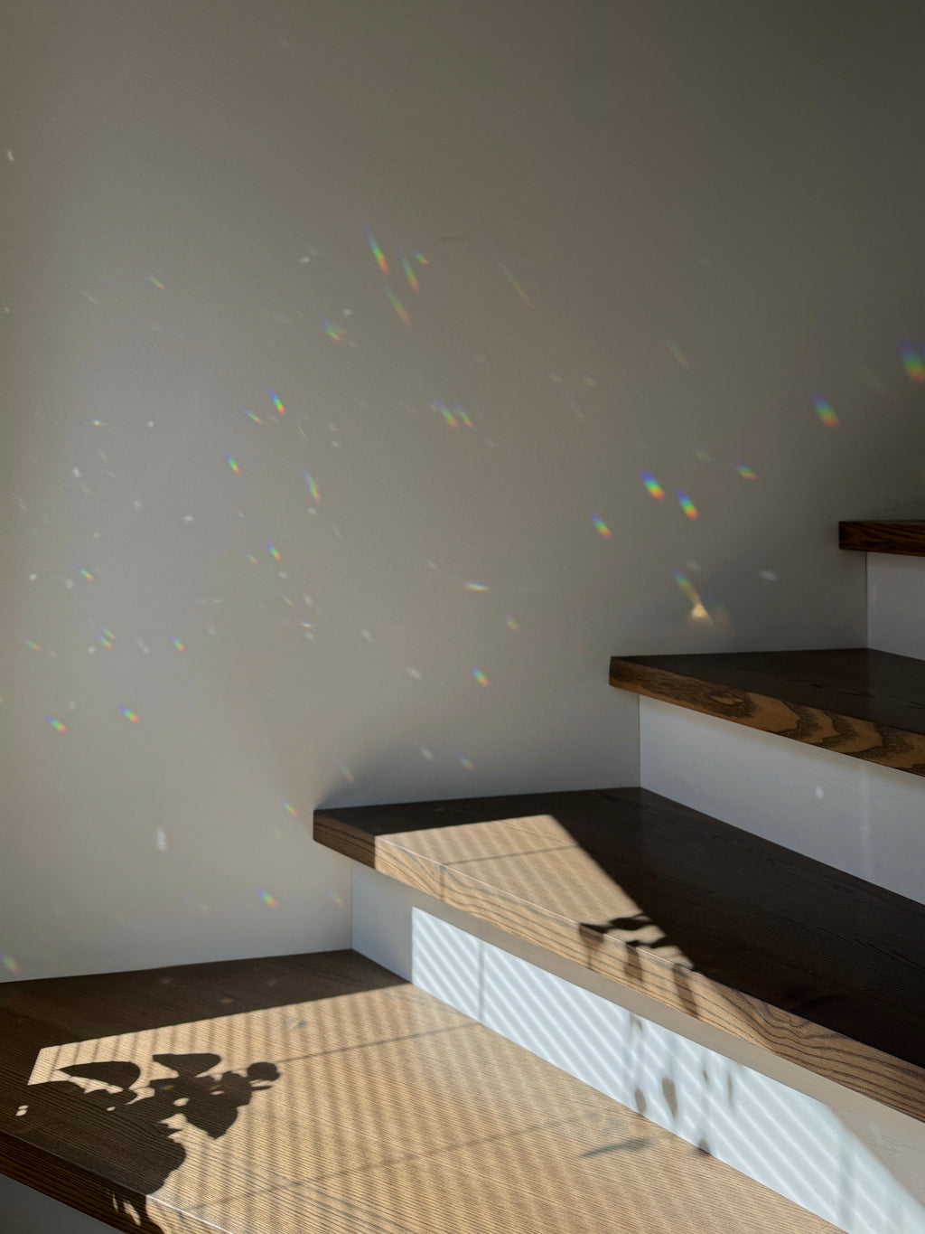 Minimalist interior corner with dark wood steps and a light beige wall, featuring natural sunlight casting striped shadows and colorful prism-like light flecks across the scene.