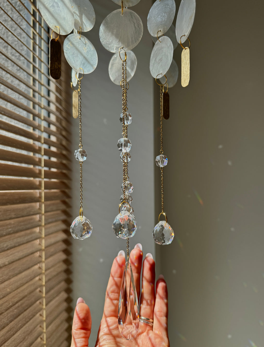 A hanging decorative wind chime with oval white shell-like discs, gold-toned chains, clear faceted crystal drops, and small gold and brown rectangular charms, shown in front of window blinds with a person's hand reaching toward it.