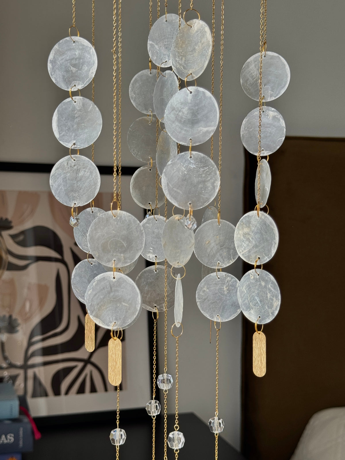A hanging wall ornament or chandelier made of multiple translucent round discs suspended on gold chains, with a few clear crystal pendants and small gold rectangular tags, shown indoors against a neutral wall.