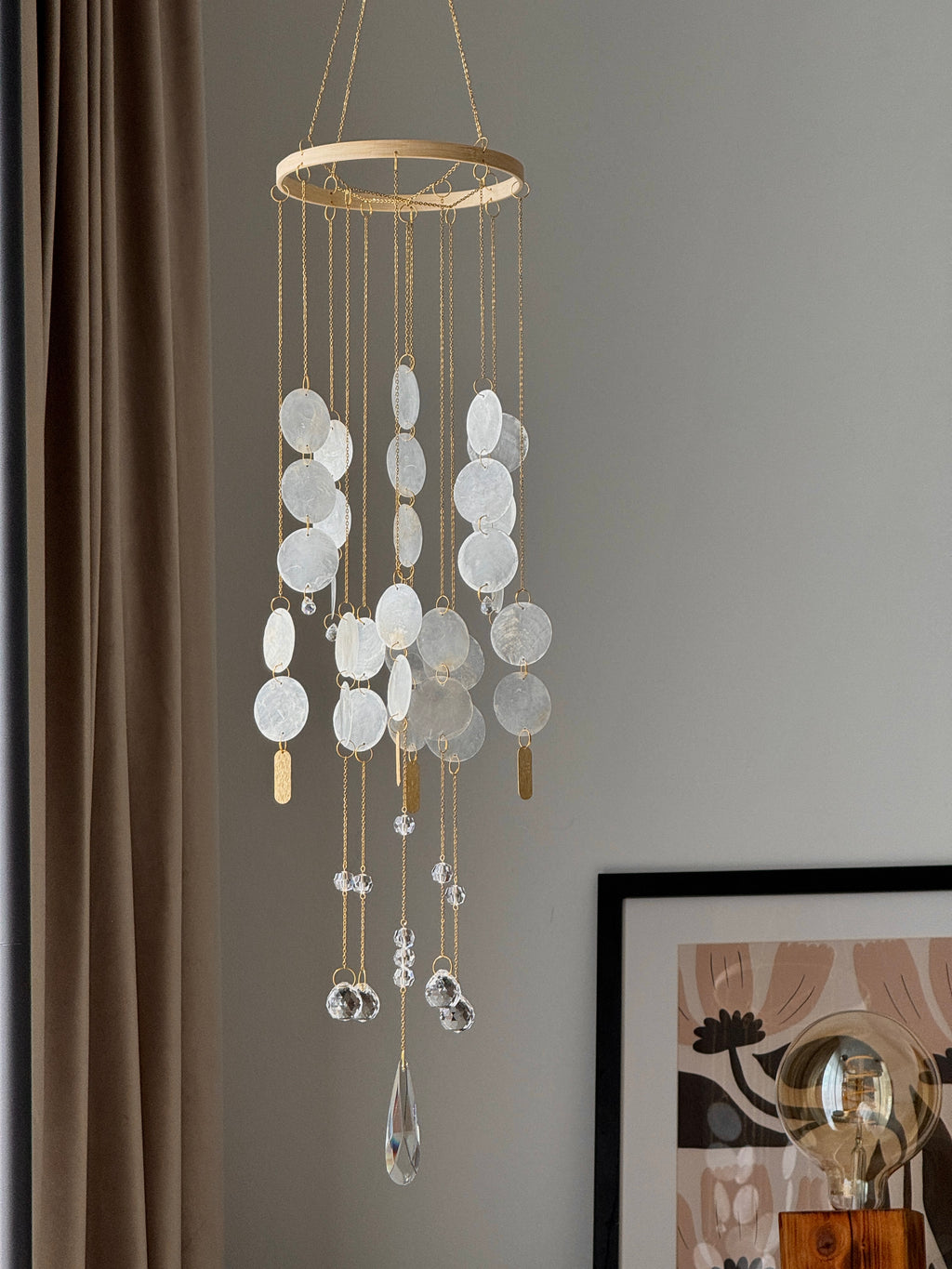 A hanging wind chime ornament with a circular gold frame, clear textured disc pendants, and dangling clear crystal teardrop beads, shown hanging in a room next to a brown curtain and framed wall art.
