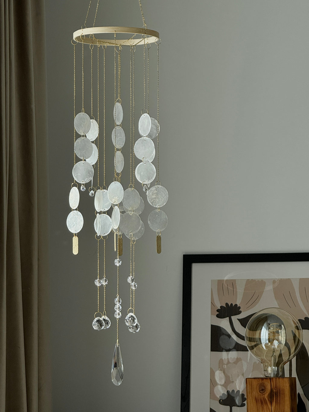 A hanging wind chime ornament with a circular gold-toned top ring, multiple strands of white disc shells, and clear teardrop glass pendants, shown against a gray wall in a styled interior setting.