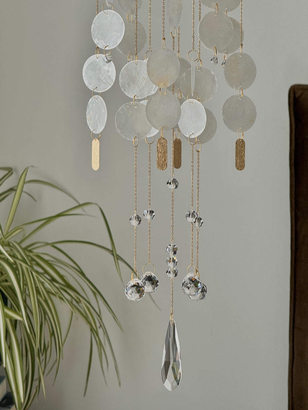 A bohemian-style hanging wind chime with translucent white disc shells, gold-toned chains, textured gold rectangular charms, and clear faceted crystal balls and teardrop pendants, shown hanging in a room next to a green plant.