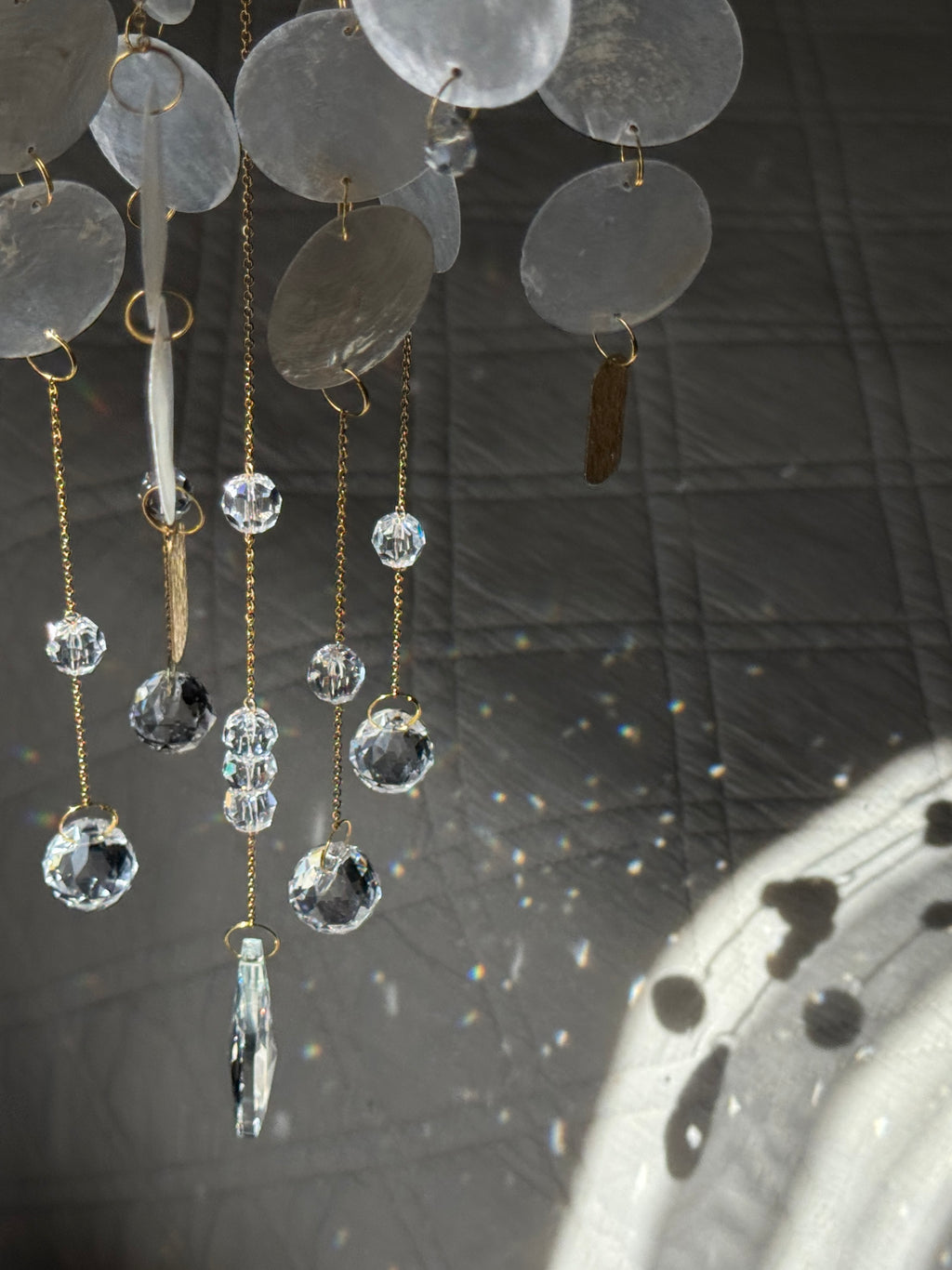 A close-up of a hanging decorative ornament or chandelier detail with translucent round discs, gold-tone chains, and clear faceted crystal beads against a dark quilted fabric background.