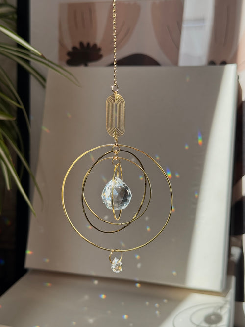 A gold-toned metal suncatcher ornament with concentric circular rings, a faceted clear crystal ball, and hanging chain, displayed in front of a light-colored panel with sunlight and rainbow reflections.