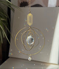 A gold-toned metal suncatcher ornament with concentric circular rings, a faceted clear crystal prism, and a hanging chain, shown in a sunlit room with rainbow light reflections on a surface.