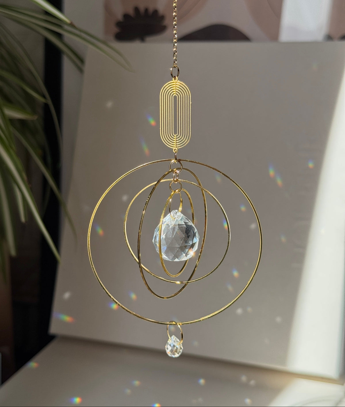 A gold-toned metal suncatcher ornament with concentric circular rings, a faceted clear crystal prism, and a hanging chain, shown in a sunlit room with rainbow light reflections on a surface.