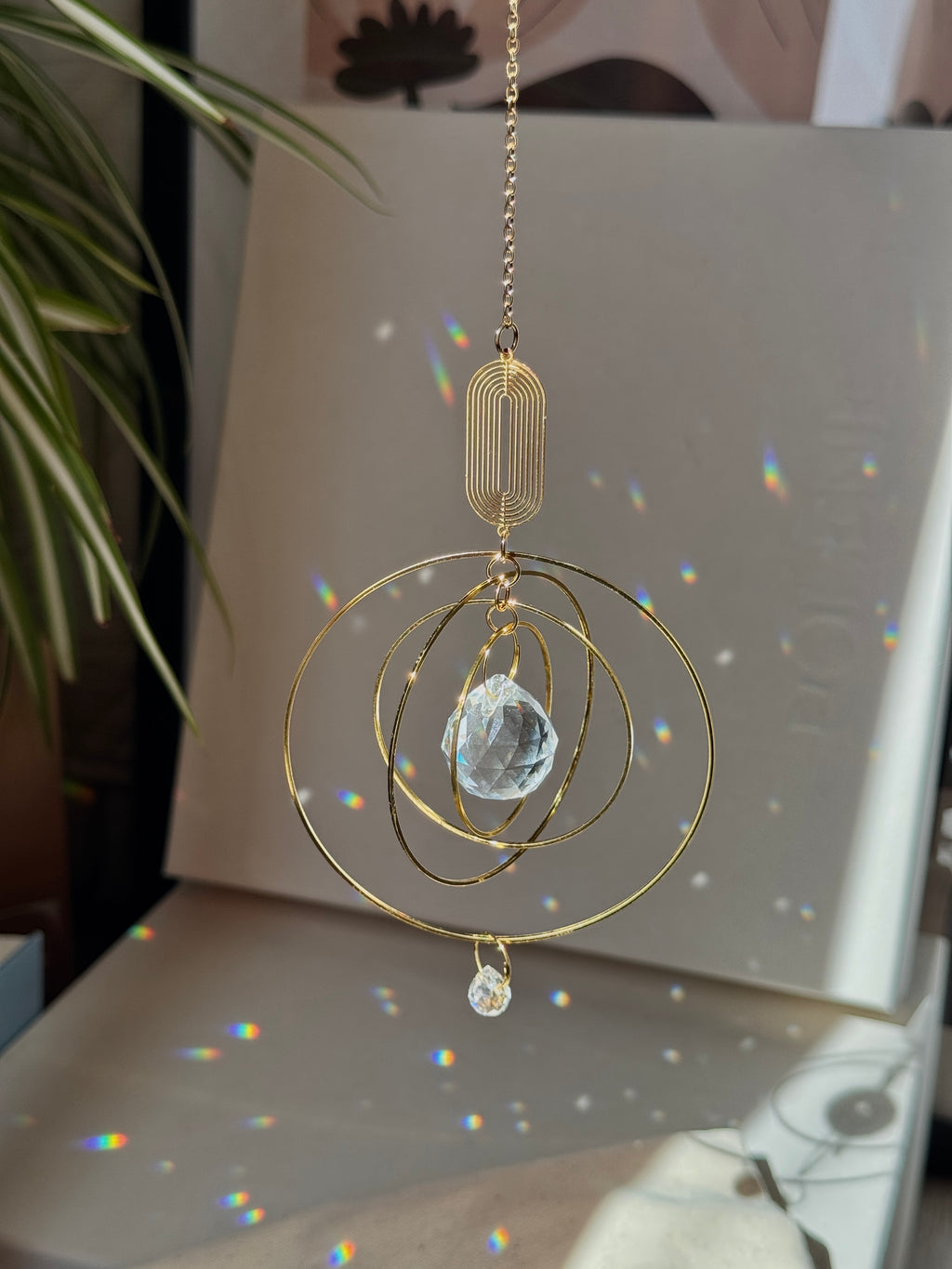 A gold-toned metal suncatcher ornament with concentric circular rings, a faceted clear crystal ball, and a small dangling teardrop charm, hanging in front of a light-colored surface with rainbow light reflections.