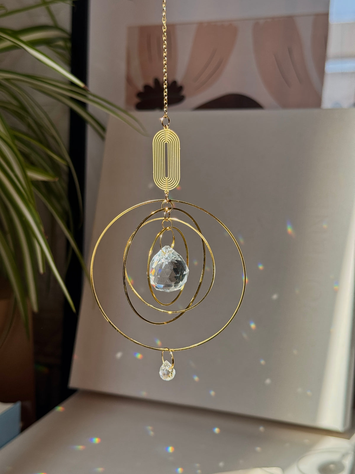 A gold-toned metal suncatcher ornament hanging from a chain, featuring concentric circular rings, a small rectangular ribbed piece, and a faceted clear crystal pendant, shown in a sunlit indoor setting with leafy plant leaves and rainbow light reflections.