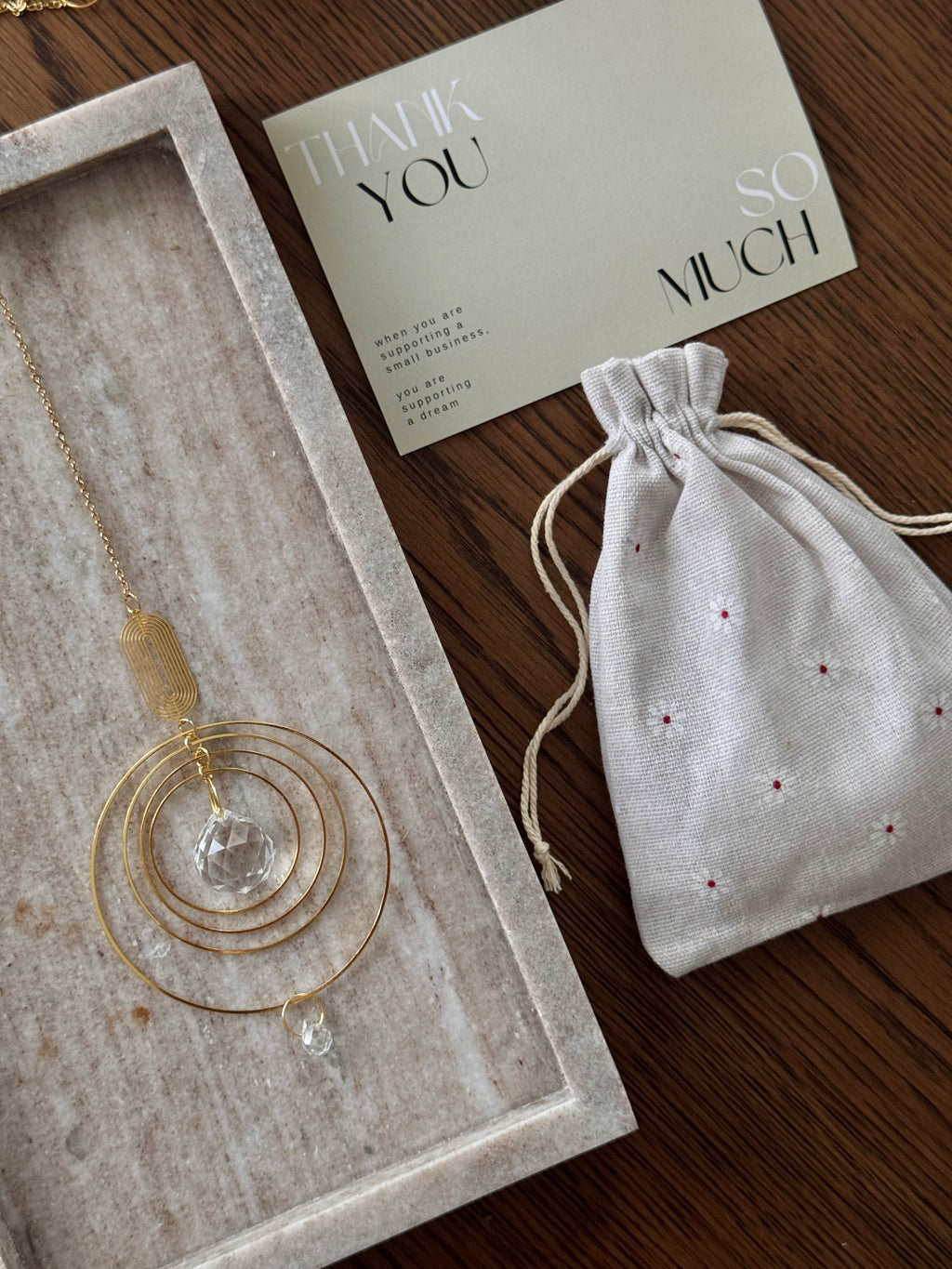 A gold-toned circular pendant necklace with a clear faceted crystal drop is displayed in a light stone tray next to a cream drawstring pouch and a thank-you card on a dark wood surface.