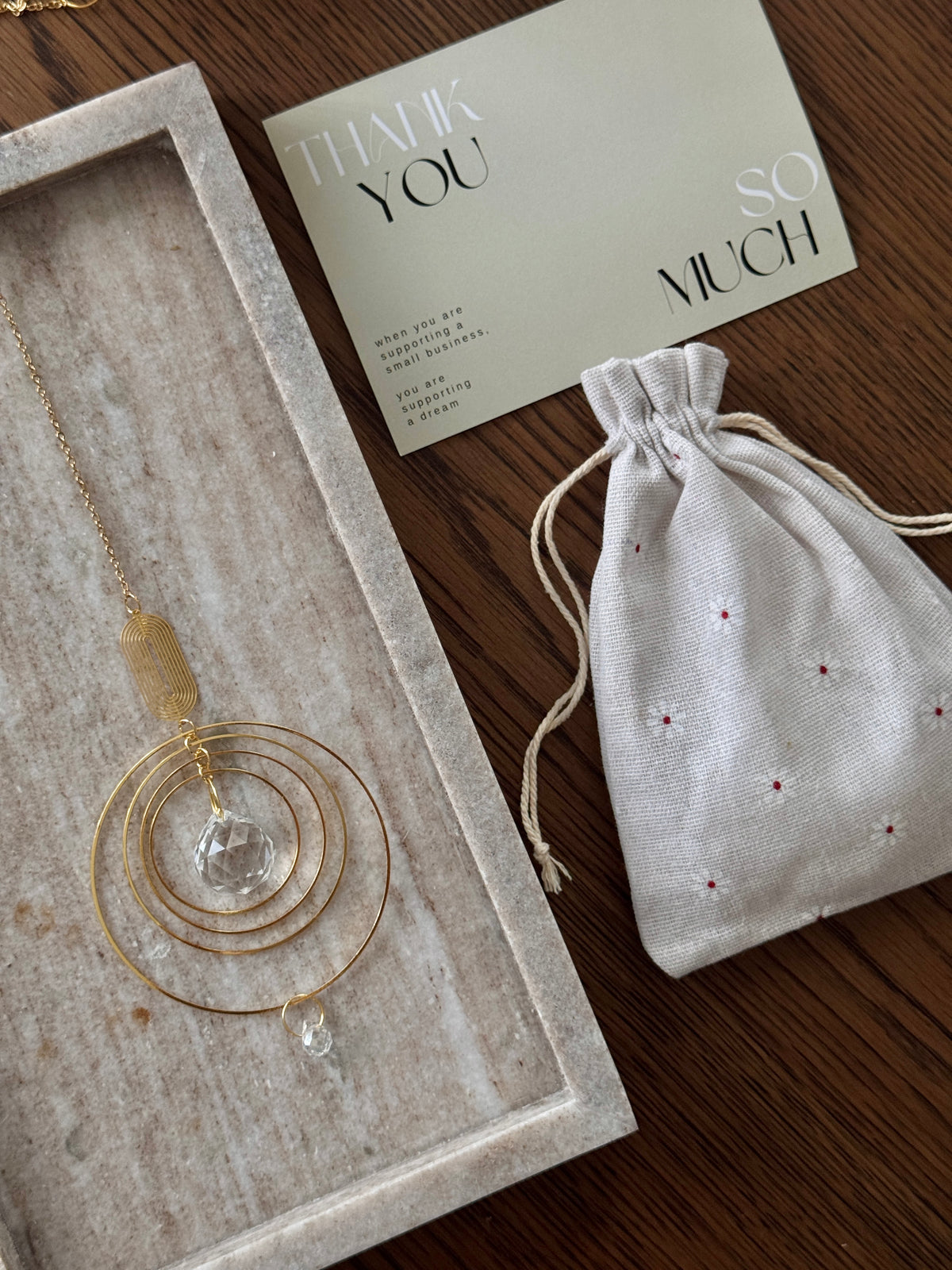 A gold-toned circular pendant necklace with a clear faceted crystal drop is displayed in a light stone tray next to a cream drawstring pouch and a thank-you card on a dark wood surface.