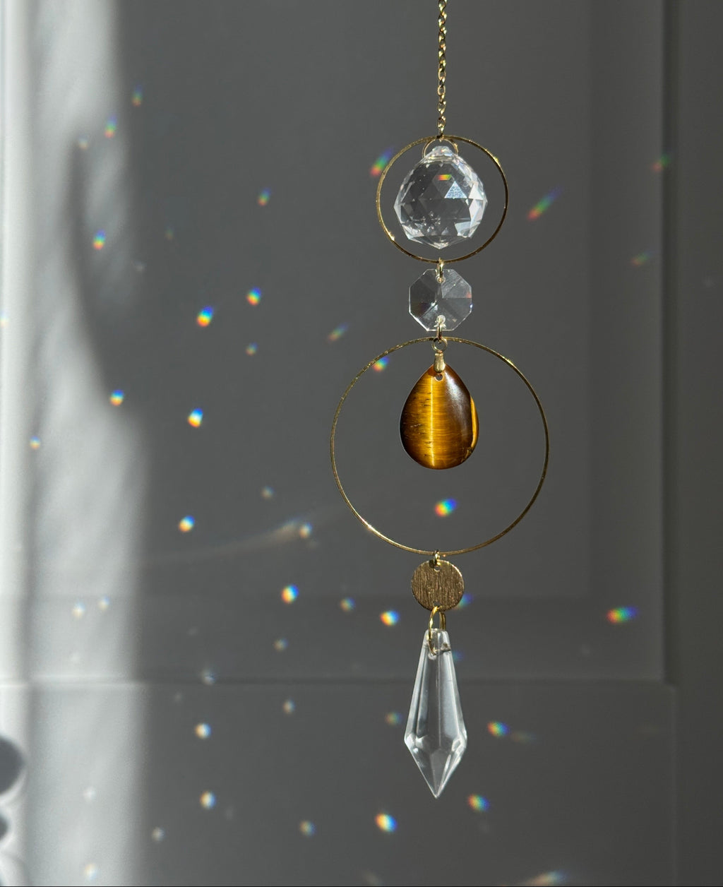 A gold-toned suncatcher with clear crystal and amber tiger's eye elements, hanging in front of a window with light and rainbow reflections.