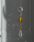 A gold-toned suncatcher with clear crystal and amber tiger's eye elements, hanging in front of a window with light and rainbow reflections.