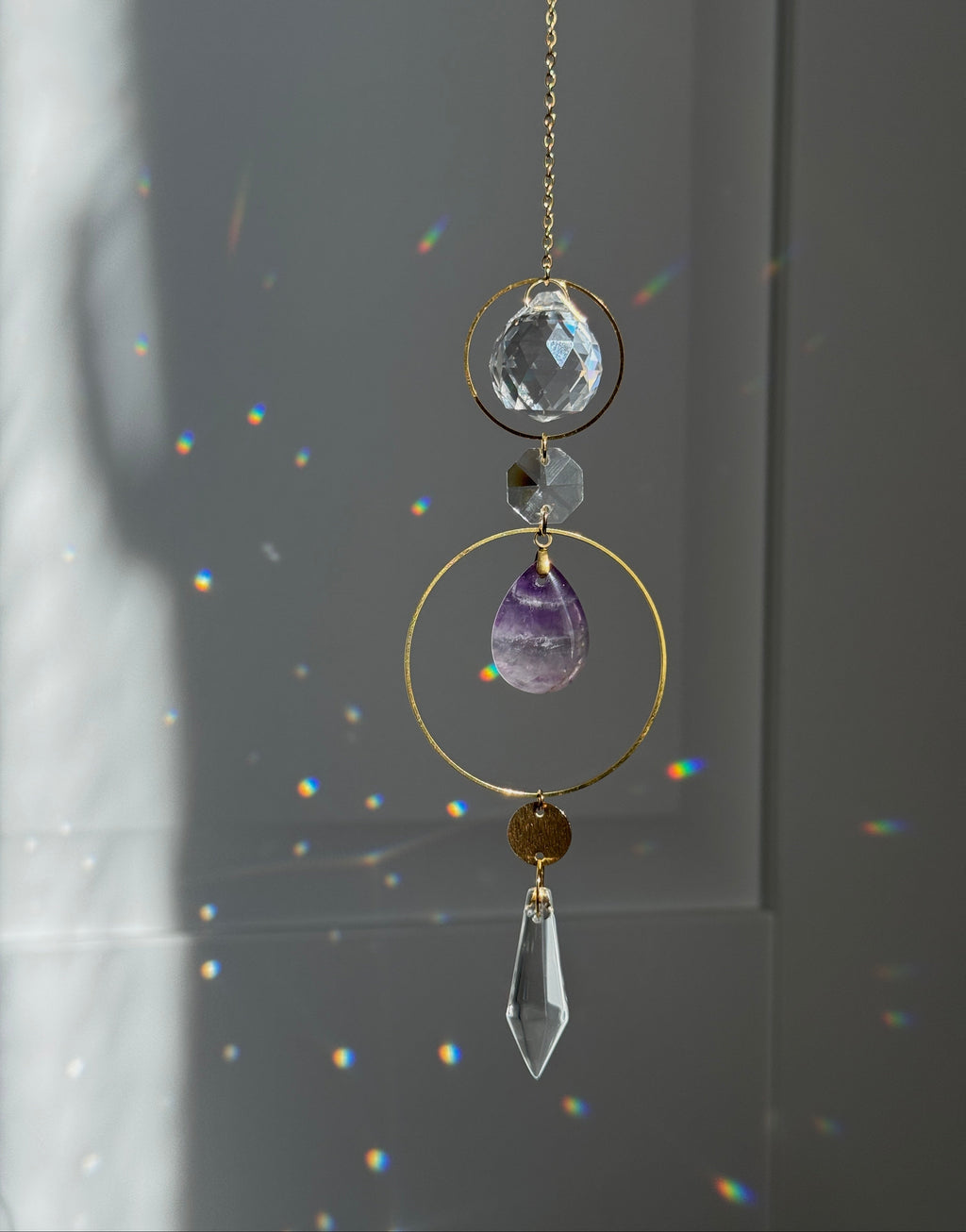 A gold suncatcher charm with clear crystal and purple amethyst elements, hanging in front of a window with sunlight and rainbow light leaks.