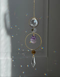A gold suncatcher charm with clear crystal and purple amethyst elements, hanging in front of a window with sunlight and rainbow light leaks.