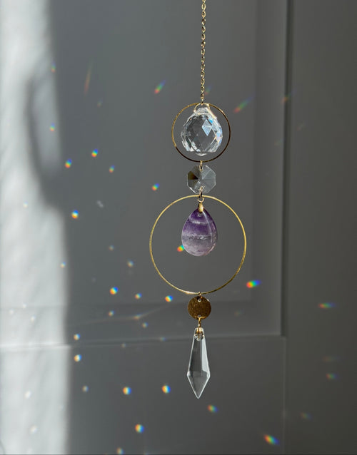 A gold suncatcher charm with clear crystal and purple amethyst elements, hanging in front of a window with sunlight and rainbow light leaks.