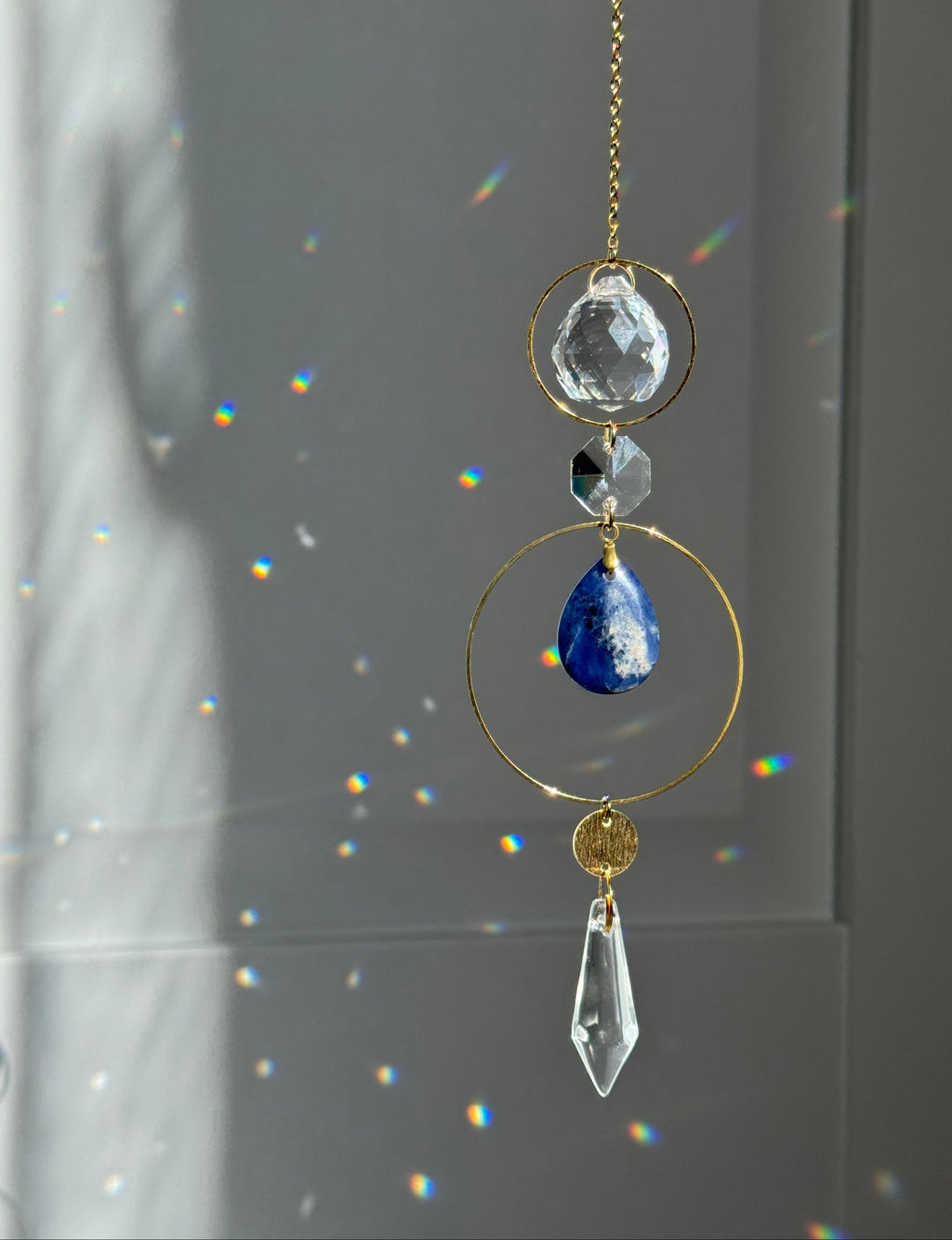 A gold suncatcher ornament hanging in front of a window, featuring a faceted clear crystal ball, a small dark faceted bead, a blue teardrop crystal, a gold ring, and a clear teardrop crystal, with rainbow light reflections on the background.