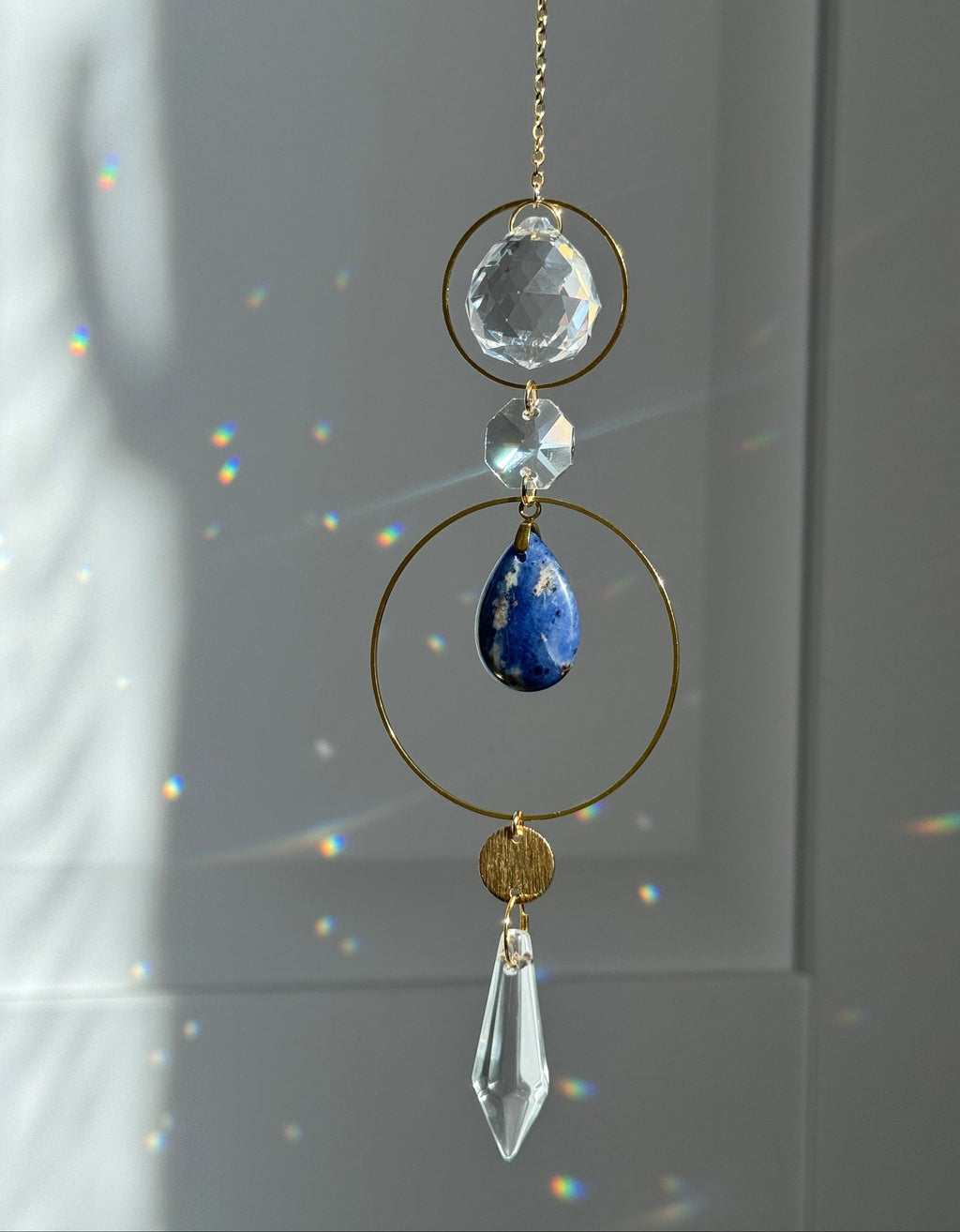 A gold suncatcher ornament with a faceted clear crystal ball, a small clear bead, a blue lapis lazuli stone, a round gold medallion, and a clear teardrop crystal pendant, hanging against a gray wall with sunlight and rainbow reflections.