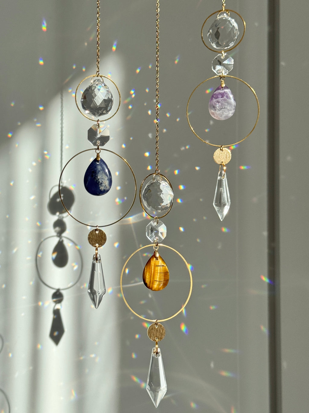 Gold circular suncatcher wind spinner ornaments with faceted crystal and gemstone beads in amber, blue, purple, and clear, hanging in front of a window with light rays and rainbow reflections.