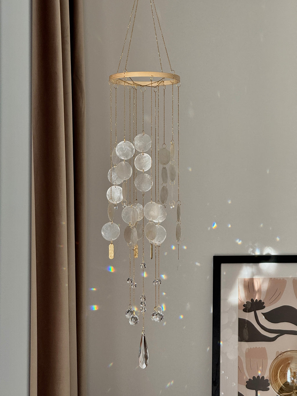 A hanging decorative wind chime with a circular wooden top, clear disc-like glass or acrylic pendants, and dangling clear crystal drops, shown hanging indoors against a light wall with curtain and framed art in the background.