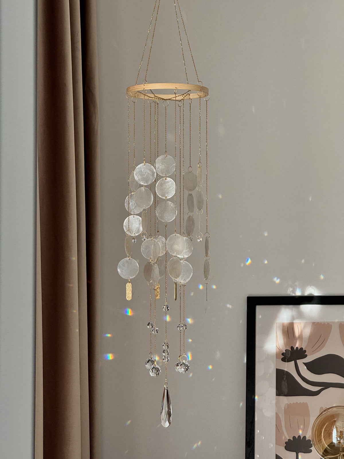 A hanging decorative wind chime with a circular wooden top, clear disc-like glass or acrylic pendants, and dangling clear crystal drops, shown hanging indoors against a light wall with curtain and framed art in the background.