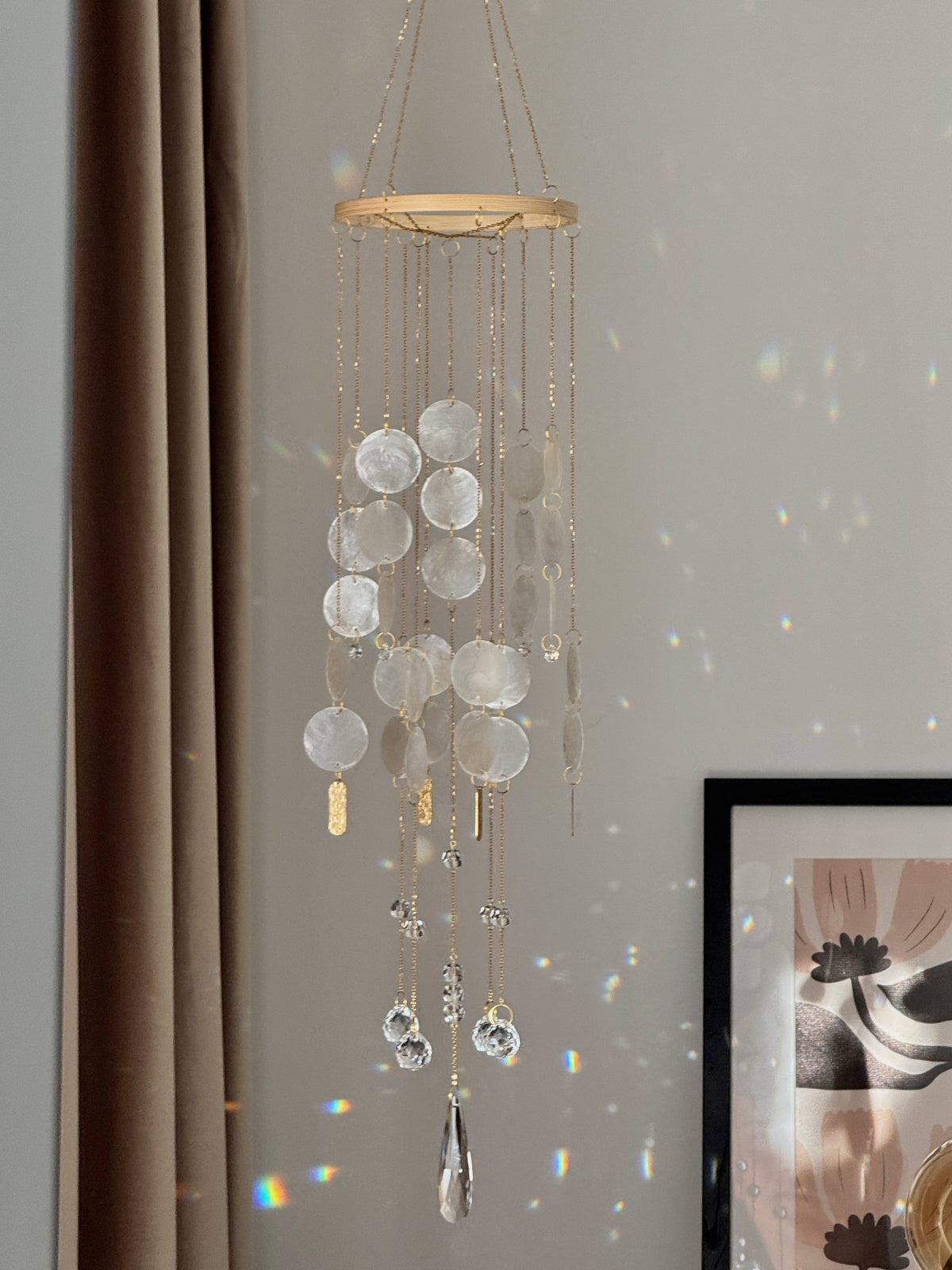 A gold-framed hanging wind chime with clear glass discs and teardrop prisms, suspended in a room with a beige wall, brown curtain, and framed art visible.
